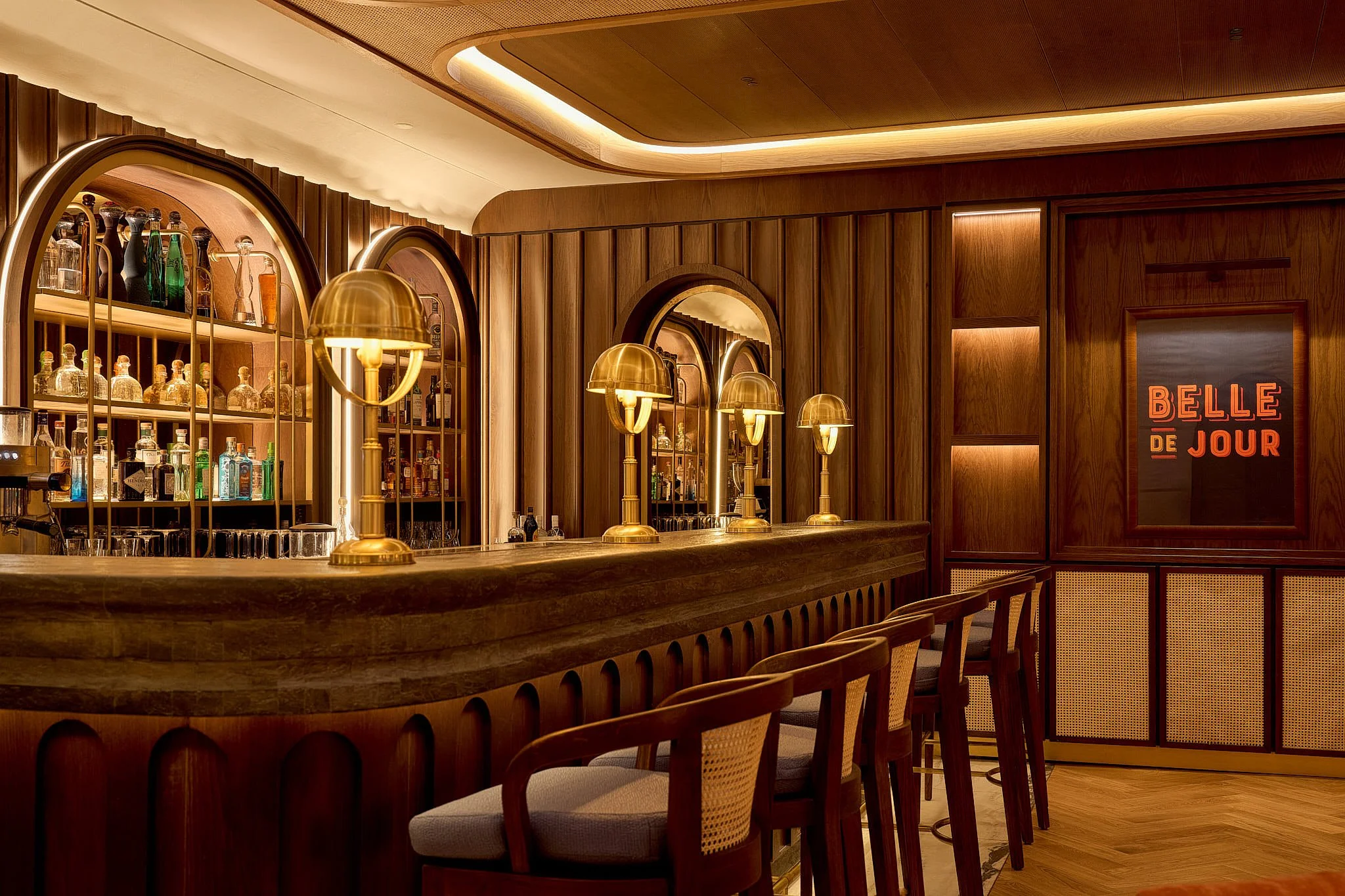 A warmly lit wooden bar with gold lamps, bottles of alcohol behind the counter, and a sign to the right that reads 'BELLE DE JOUR' in red letters.