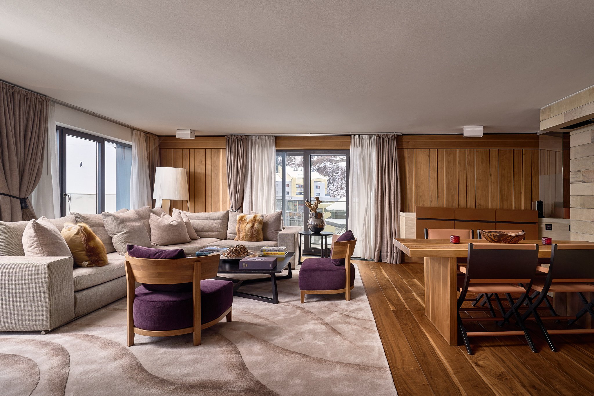 Living room with beige sectional sofa, purple armchairs, wood-paneled walls, large windows with curtains, a coffee table with books and decorative items, and a dining area with wooden table and chairs.