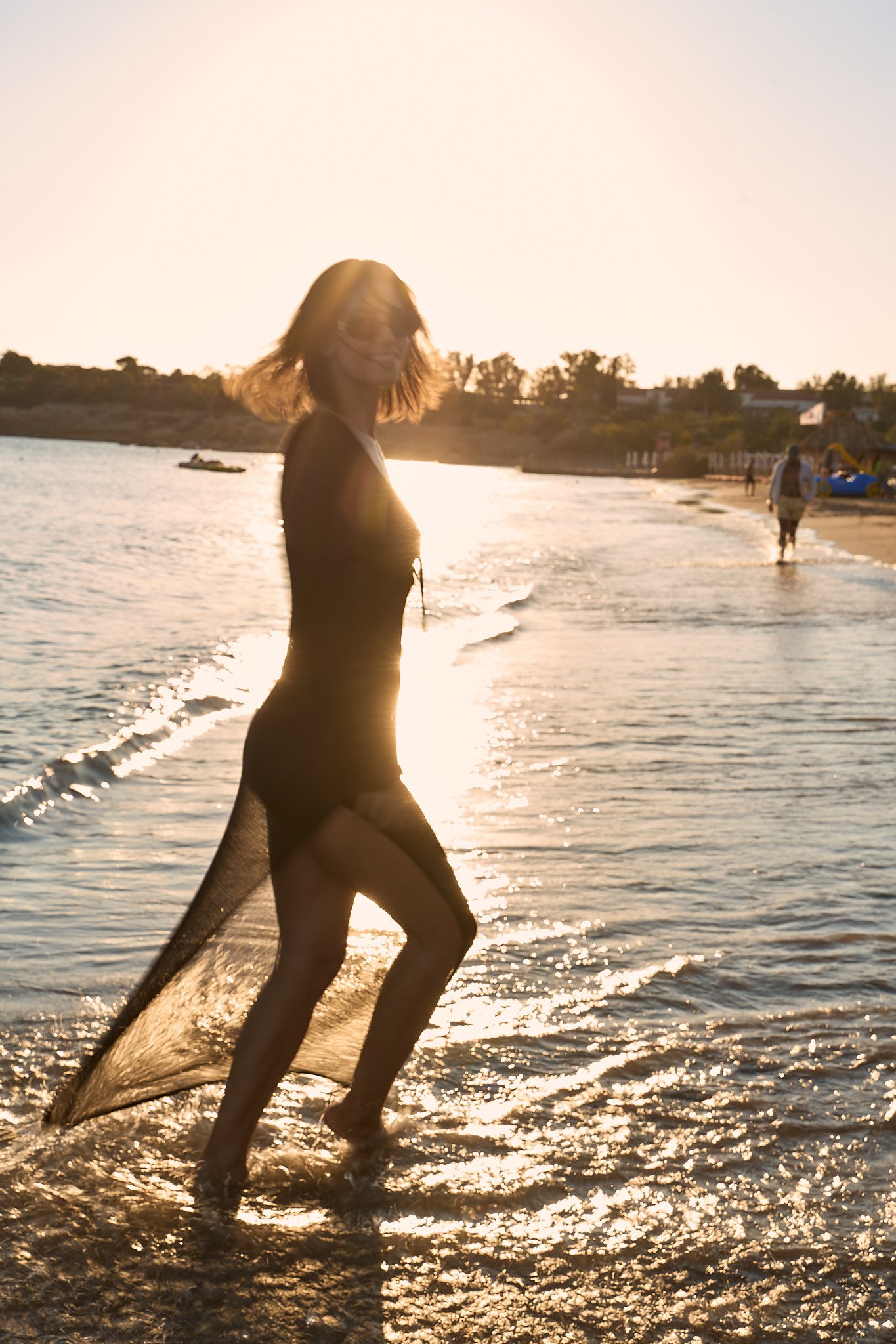 A woman in a black dress and sheer overlay walking in shallow water at sunset on a beach, with other people and boats visible in the background.