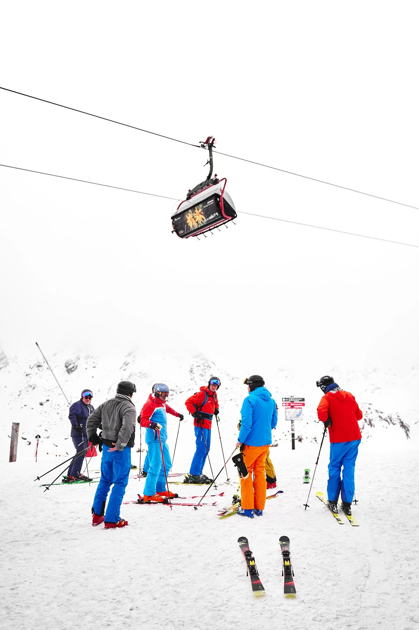 Group of skiers in colorful gear waiting on a snowy mountain, with a ski lift overhead and snow-covered peaks in the background.