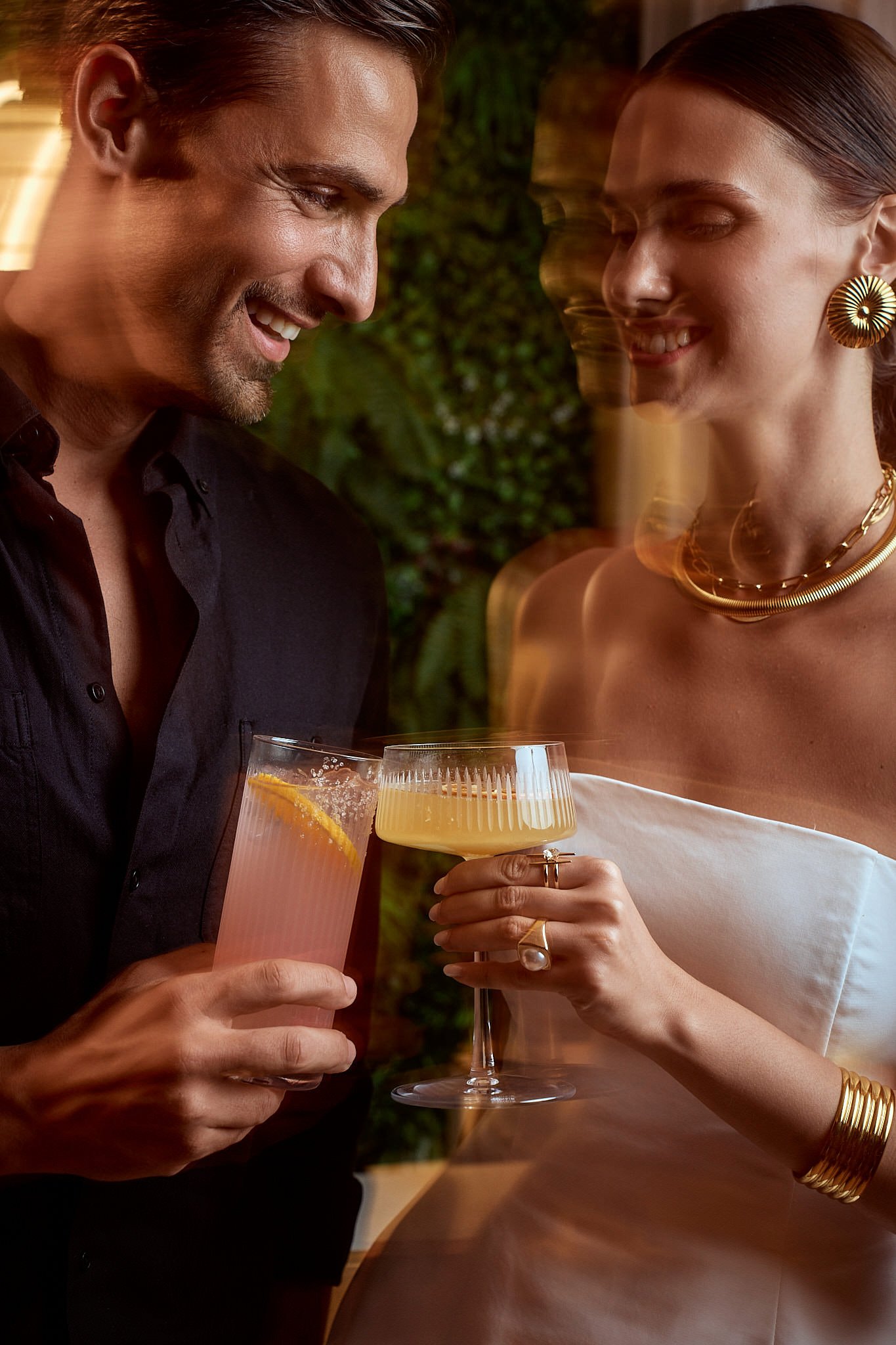 A man and a woman smiling and toasting drinks in a dimly-lit setting, with a slight motion blur effect.