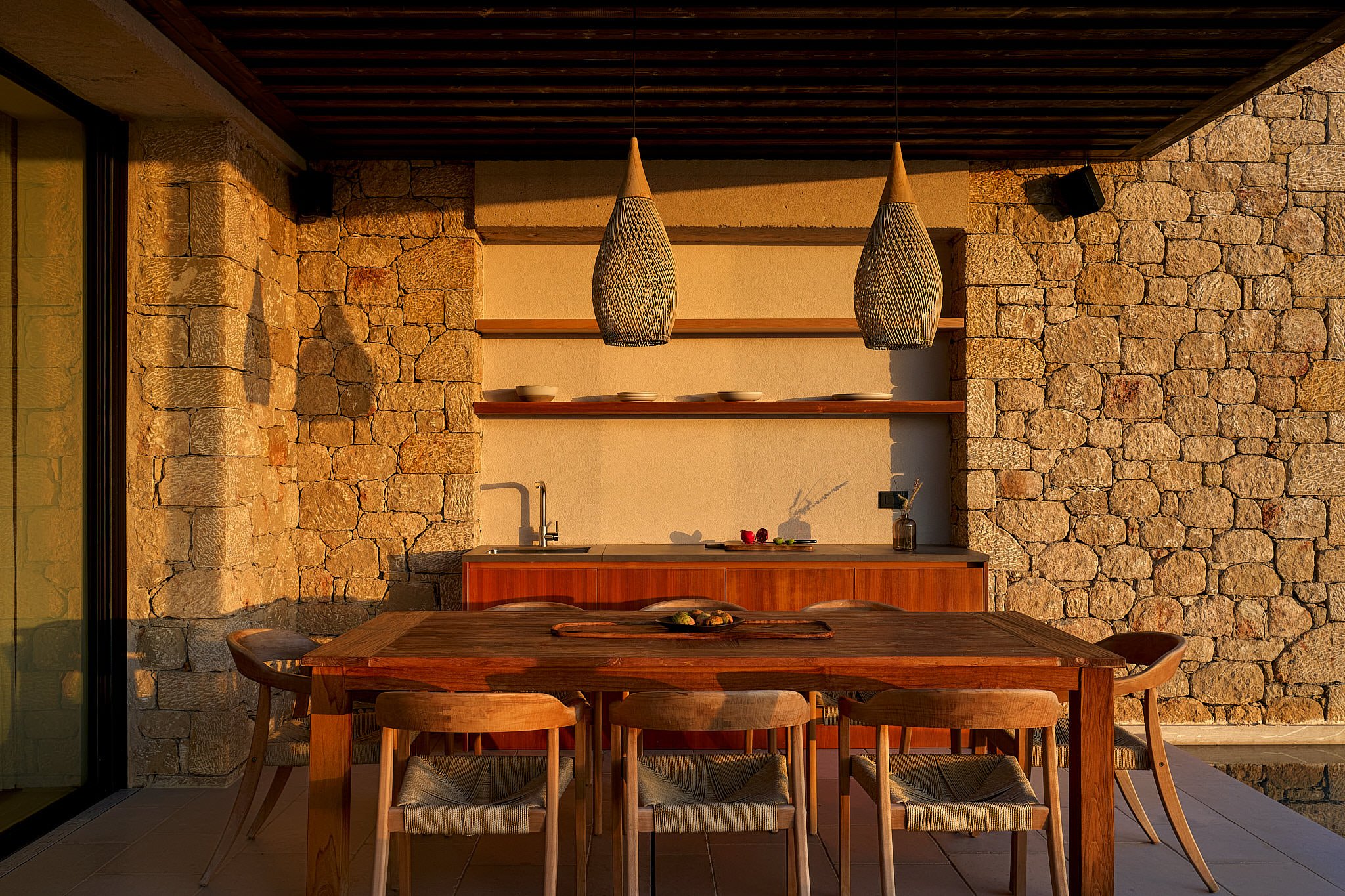 An outdoor dining area with a wooden table and six chairs, a stone wall, and two hanging pendant lights.