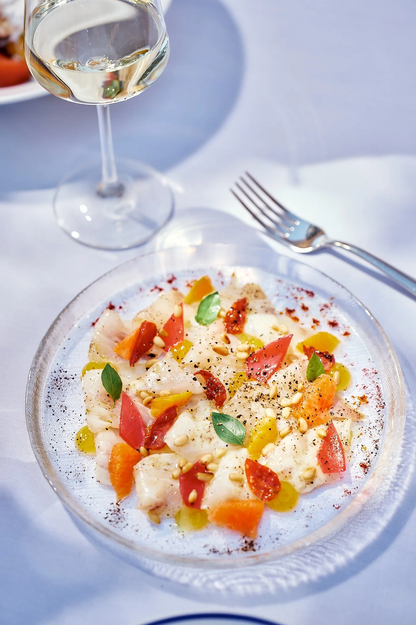 A plate of ceviche garnished with chopped tomatoes, herbs, pine nuts, and spices, accompanied by a glass of white wine on a white tablecloth.