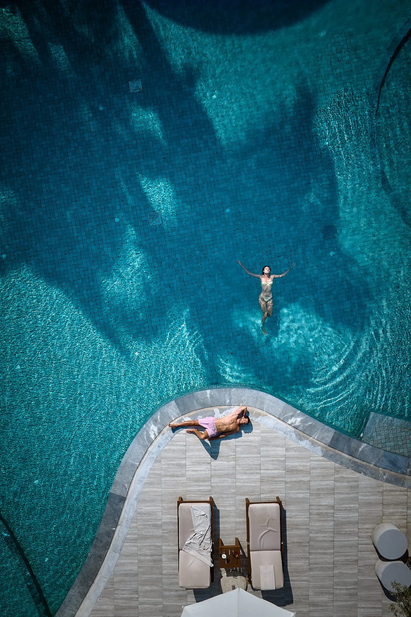 An aerial view of a swimming pool with two people relaxing and swimming, surrounded by a beige poolside area with lounge chairs and an umbrella.