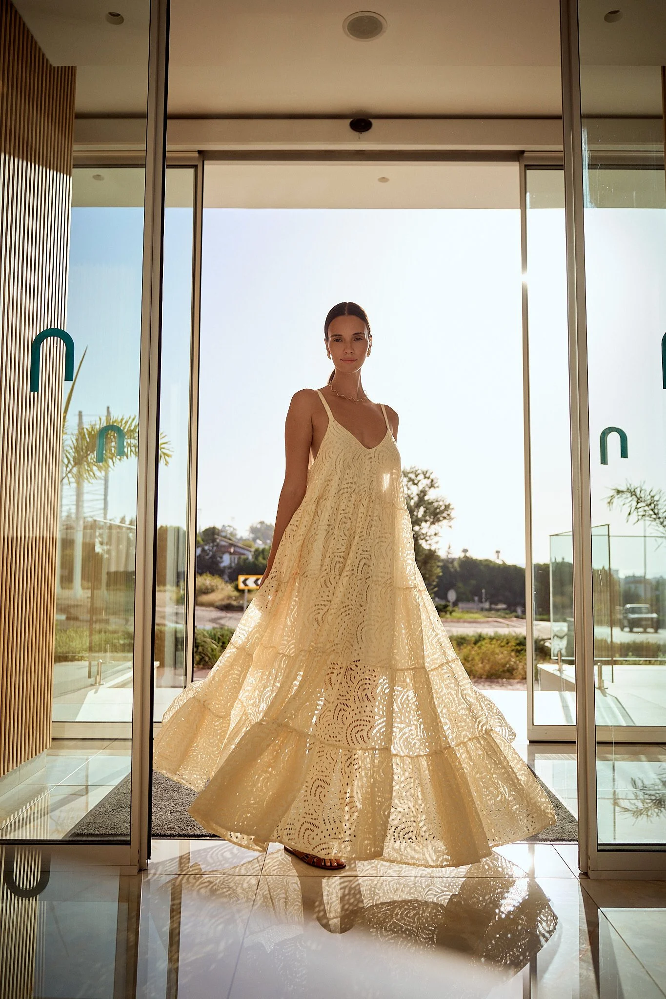 A woman in a beige, flowing lace dress standing inside near glass doors with sunlight behind her.
