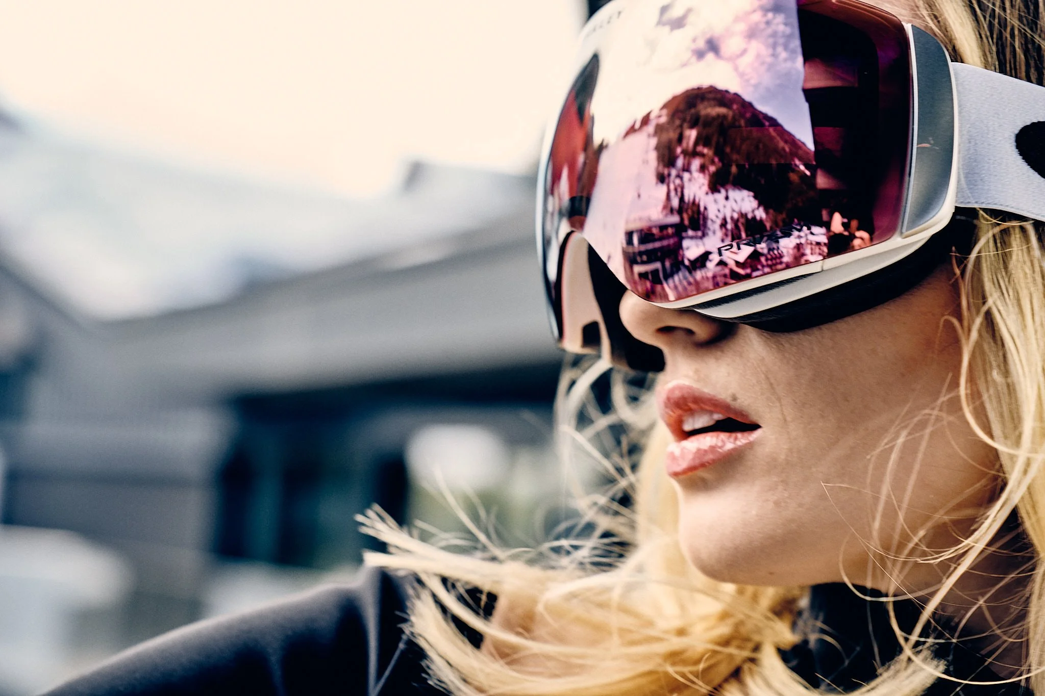 A woman wearing a snow helmet and goggles, with wind-blown blonde hair, outdoors during daytime.