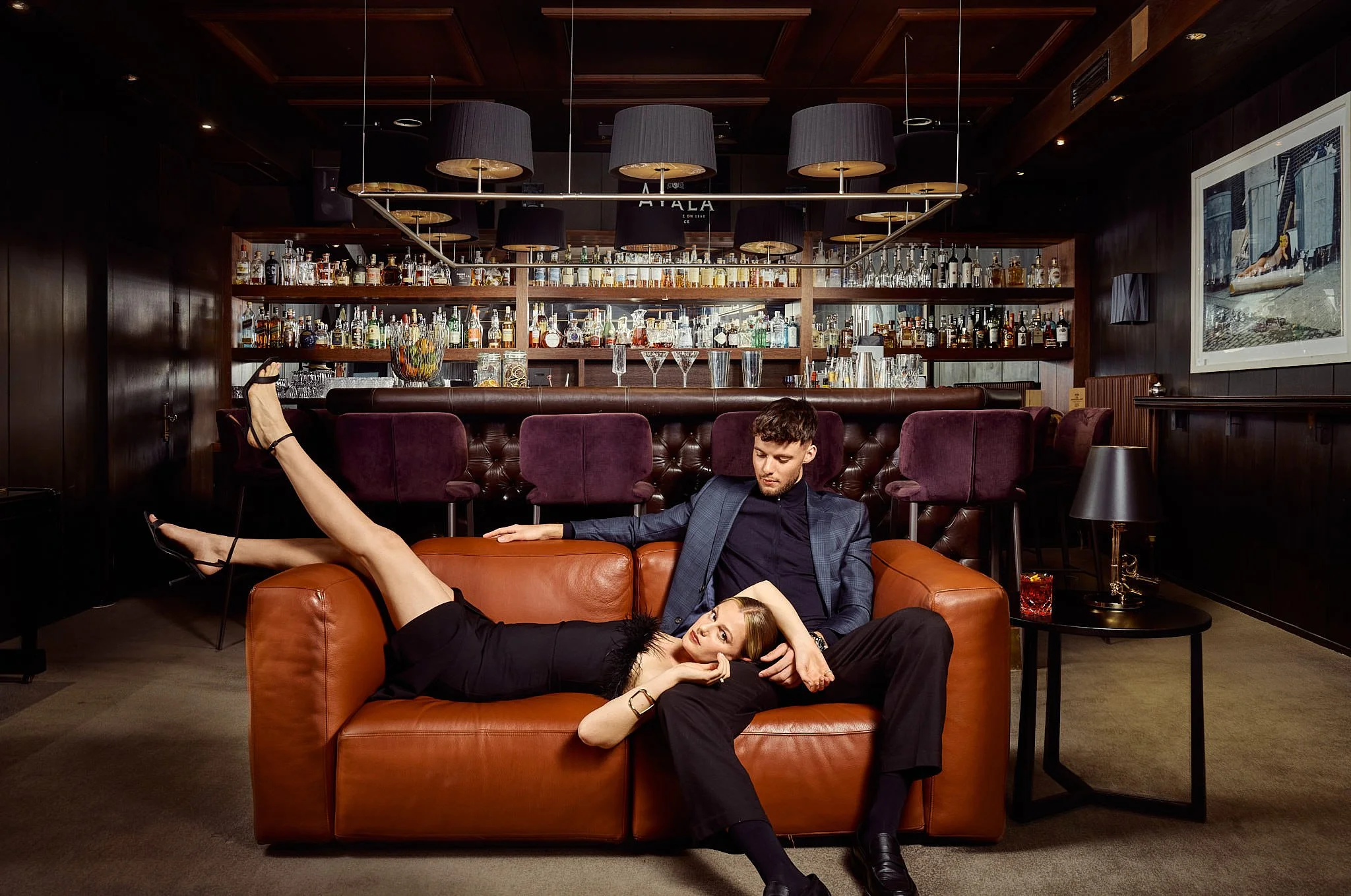 A man and woman in formal attire lounging on a brown leather couch in a dimly lit bar or lounge with a bar counter and shelves of alcohol bottles in the background.