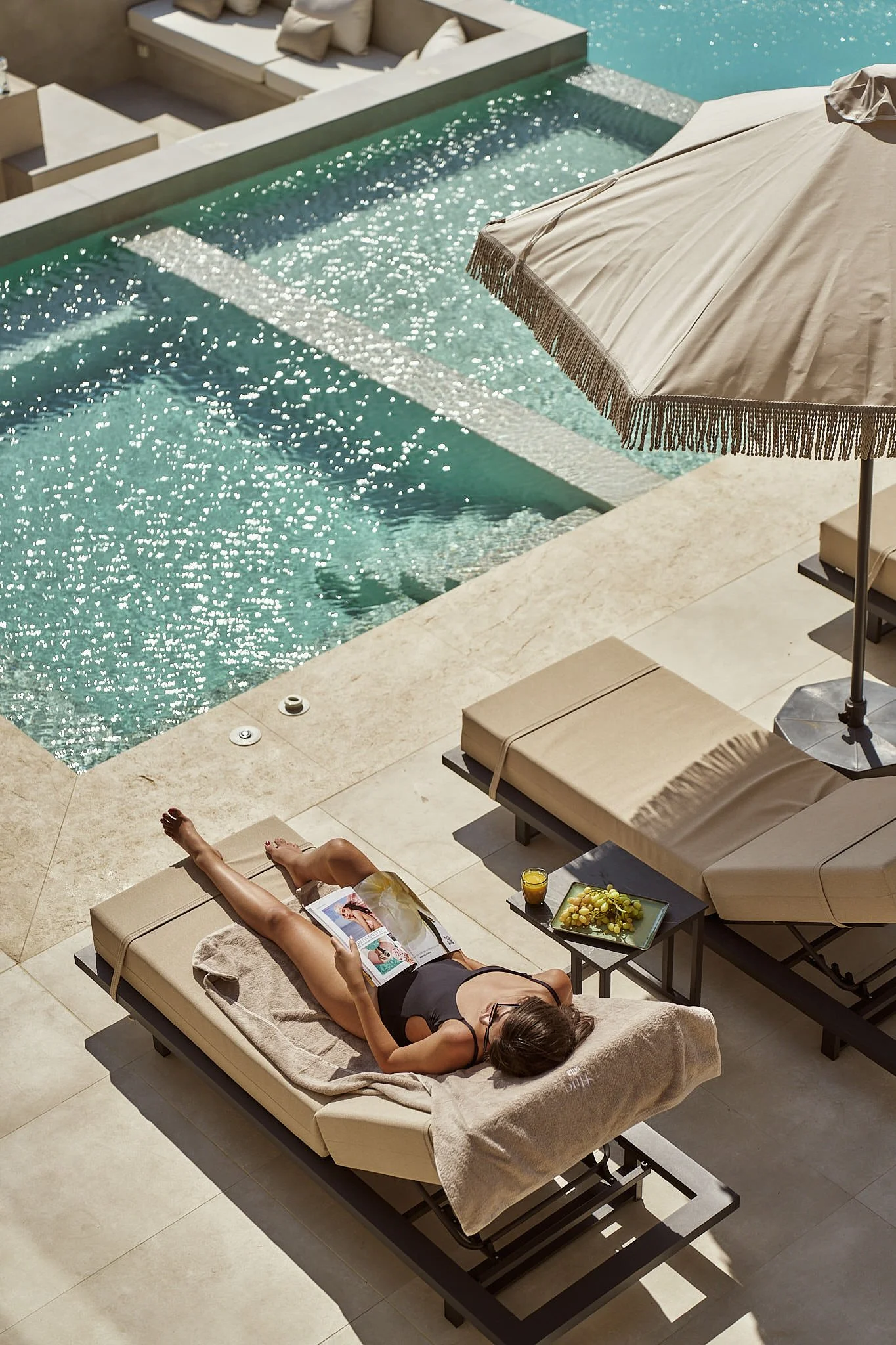 A woman lying on a lounge chair next to a swimming pool, reading a magazine. There is an umbrella providing shade and a small table with snacks and a drink.