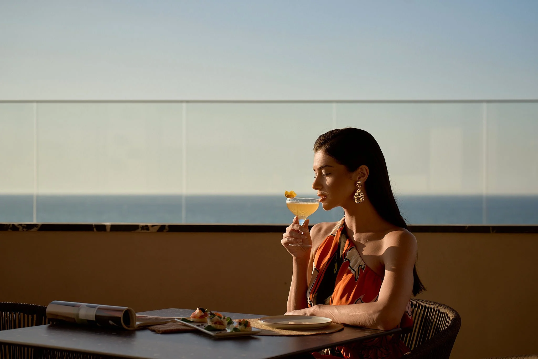Woman in a floral dress enjoying a cocktail with a pineapple garnish on a balcony overlooking the ocean.