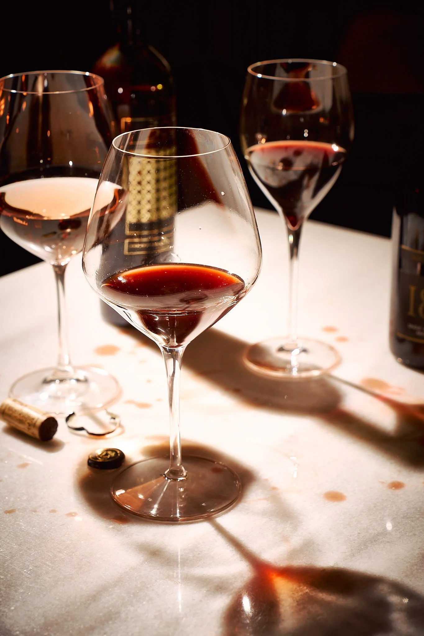 Three glasses of red wine on a white table with wine bottle and corks, shadows and wine stains.