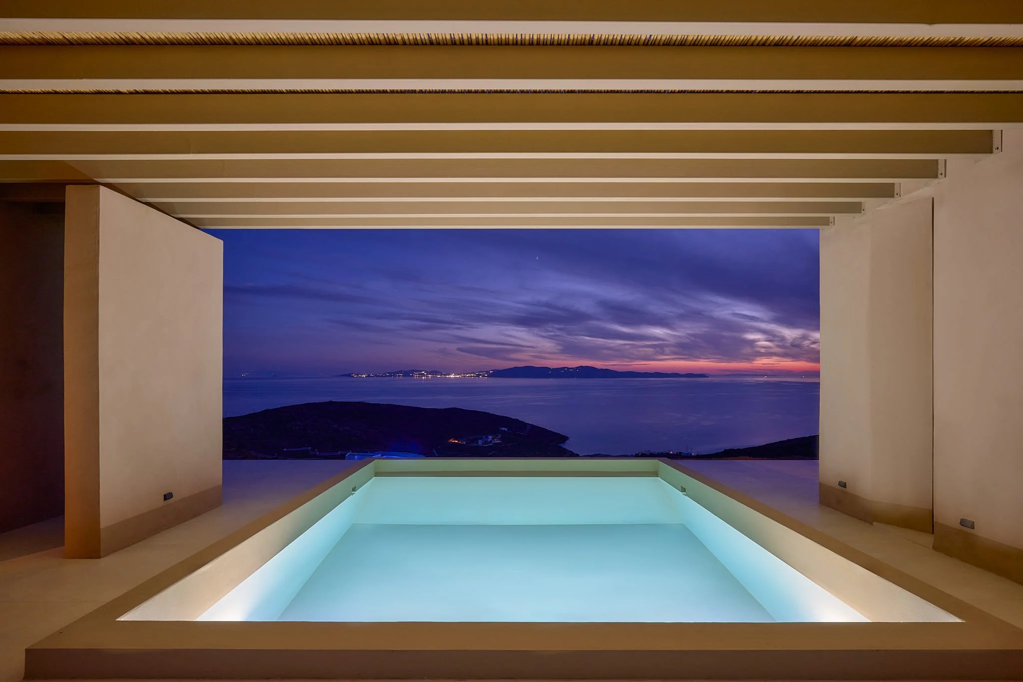 Night view from an indoor swimming pool overlooking the ocean and distant coastline, with purple and blue sky.
