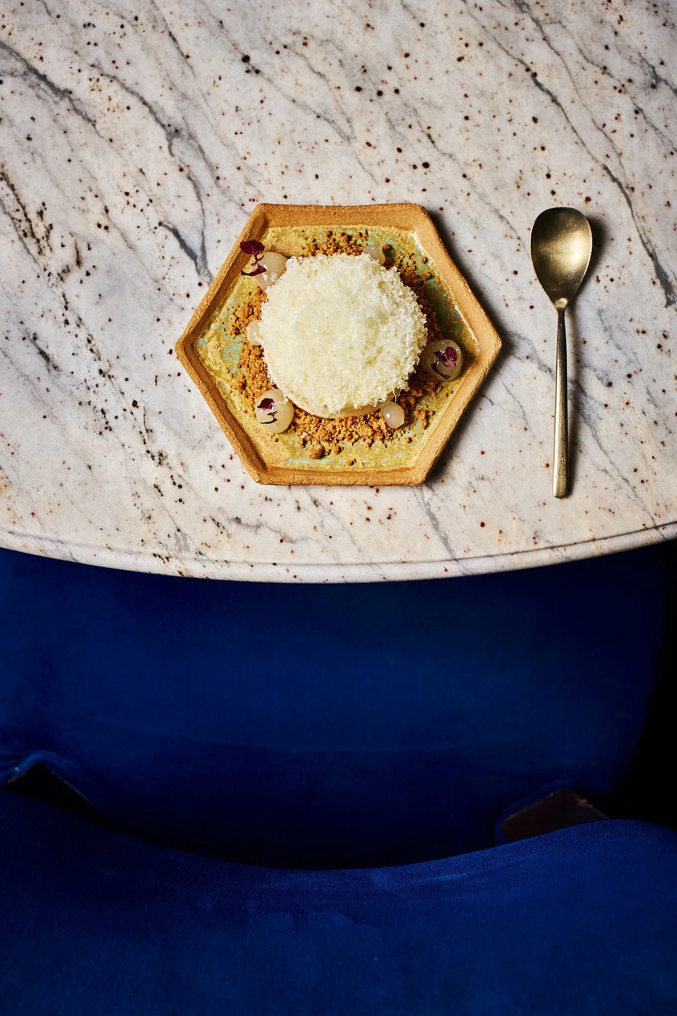 A plated dessert on a hexagonal dish with a white cream or mousse topped with coconut flakes, garnished with small edible flowers and spheres, on a marble table with a gold spoon nearby.