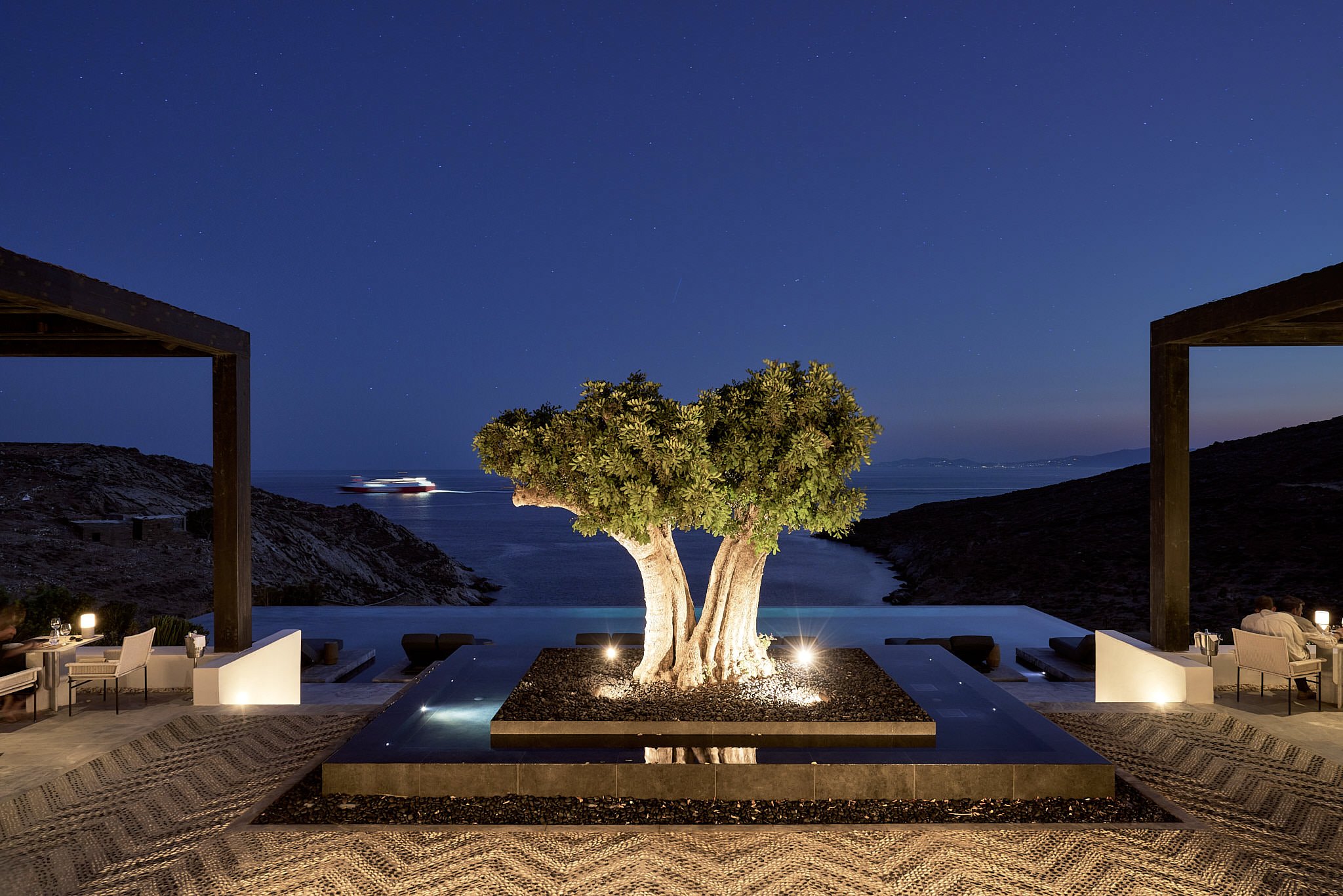 Night view of an outdoor dining area with a central illuminated tree, overlooking the ocean and a cruise ship in the distance, framed by two dark structures.