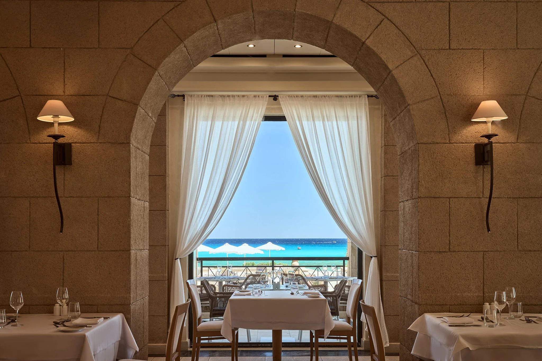 Indoor restaurant with stone archway and wall sconces, opening to a balcony with white tablecloths, glassware, and overlooking a beach with umbrellas and ocean.