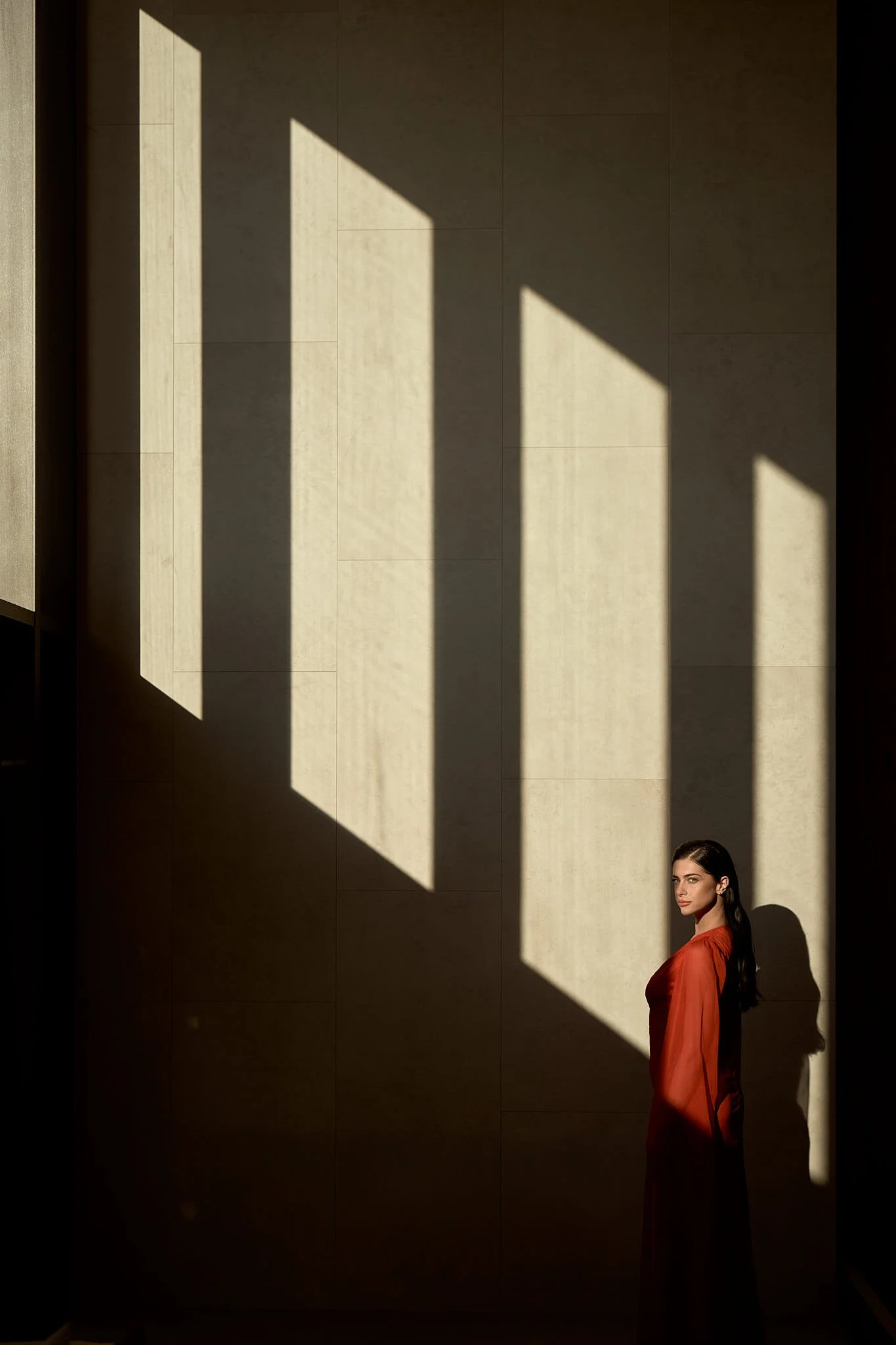 A woman with dark hair in a red dress standing in shadows with geometric window shadows projected on a beige wall.