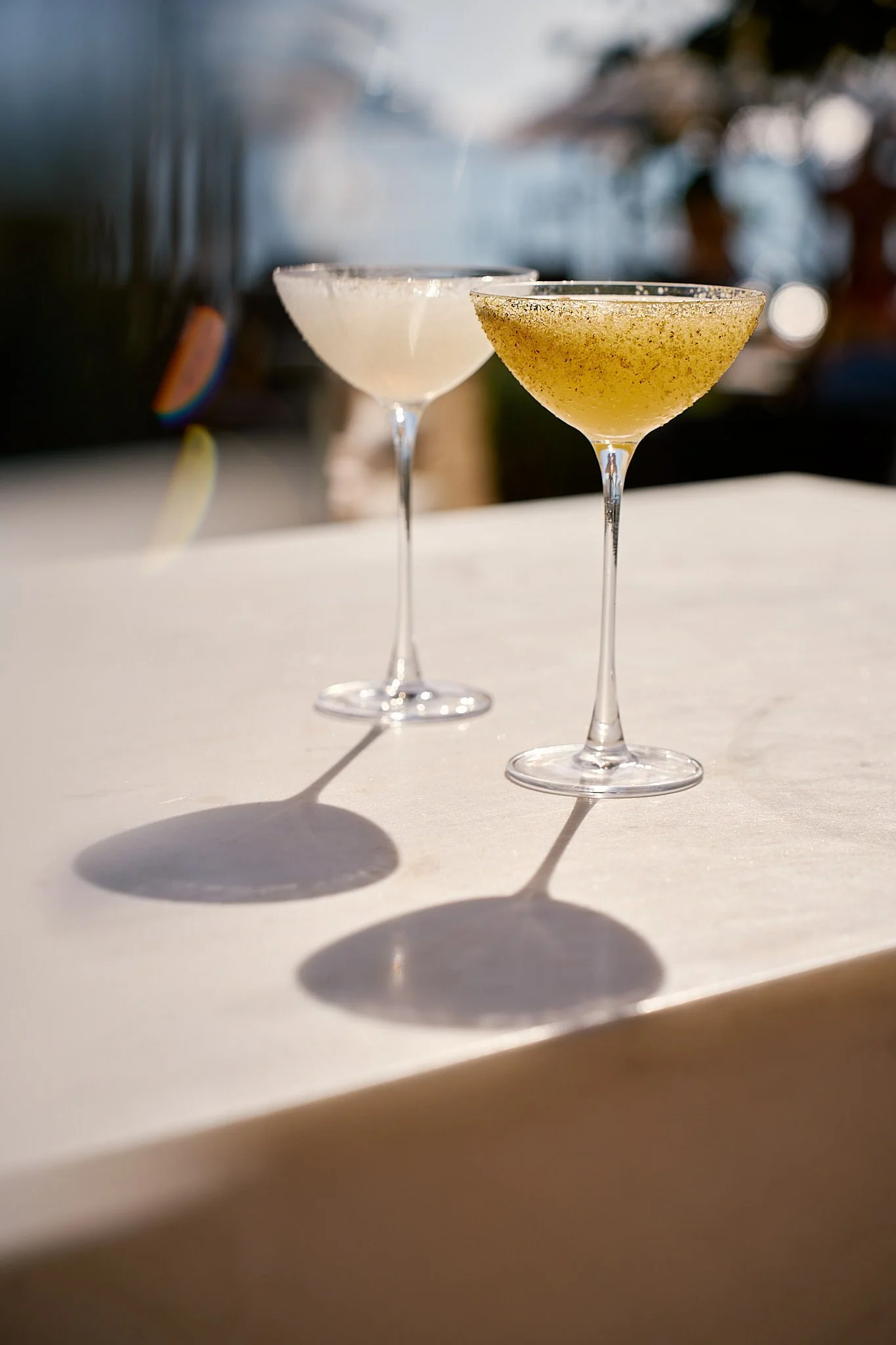 Two cocktail glasses on a white surface, casting shadows in sunlight, with a blurred outdoor background.