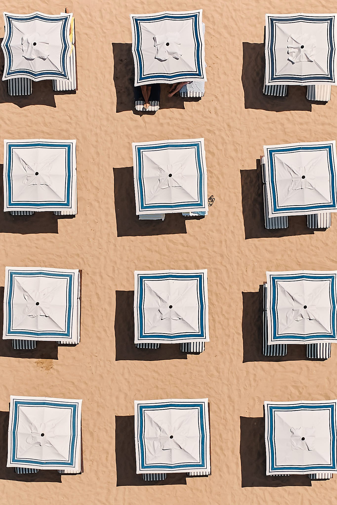 Aerial view of beach umbrellas arranged in a grid on sandy beach, with shadows cast to the side.