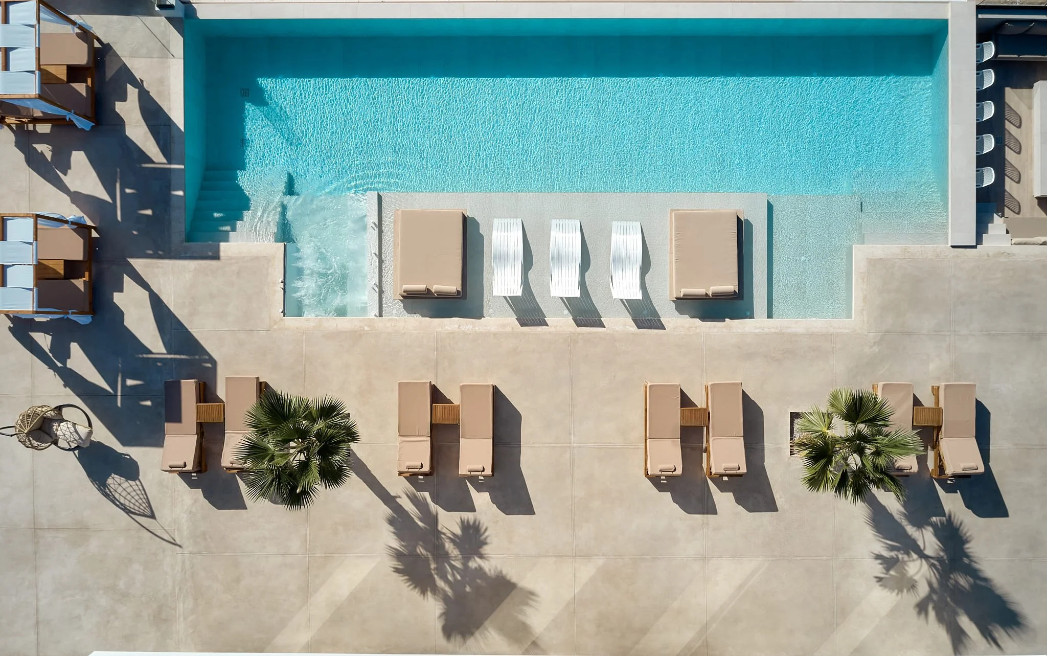 Aerial view of a swimming pool with sun loungers, palm trees, and shaded seating areas around it