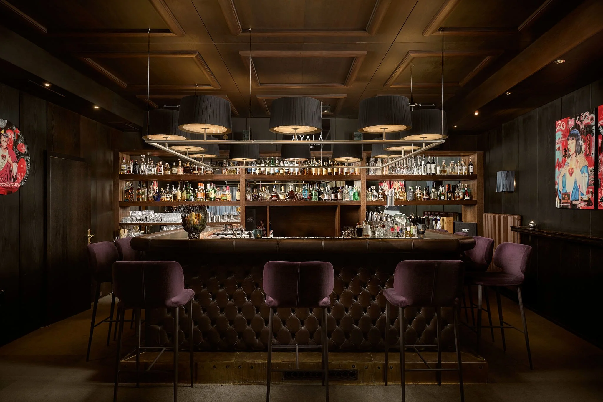 Dark wooden bar with purple velvet chairs, illuminated by black pendant lamps, stocked with various bottles of alcohol and glassware, with dark wood-paneled walls and colorful artwork.