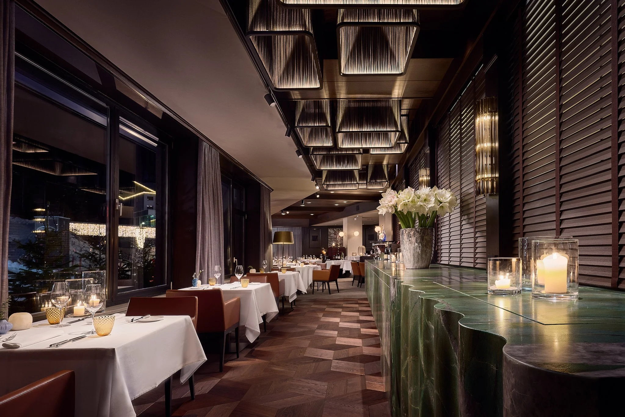 An elegant restaurant interior with large windows, white tablecloths, wine glasses, candles, and a green marble bar adorned with a vase of white flowers.