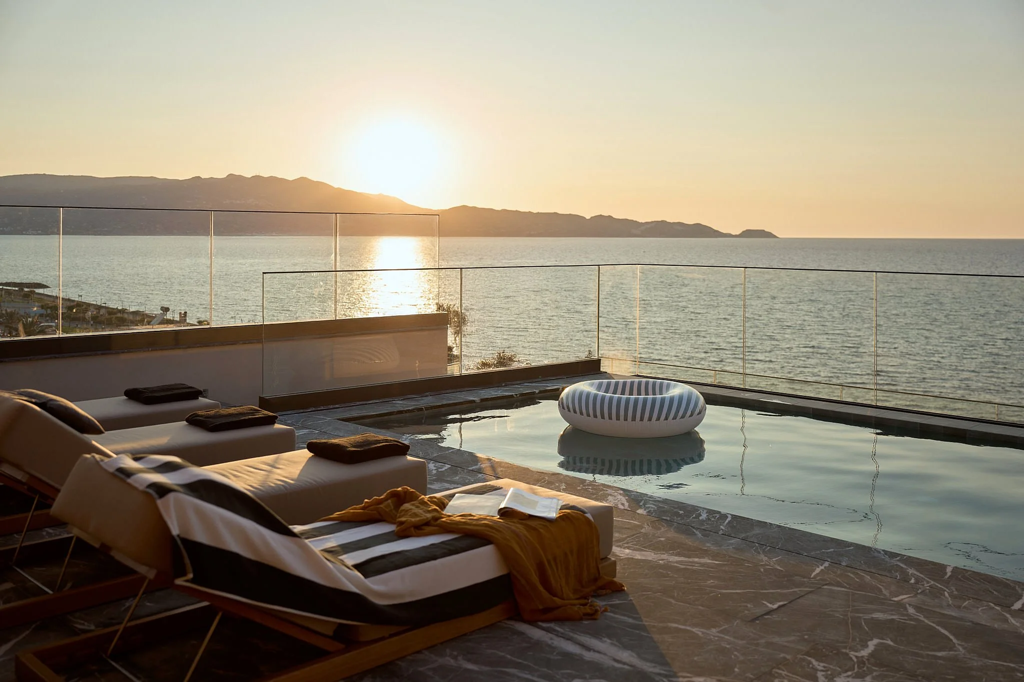Luxury outdoor pool area with lounge chairs, towels, a book, and a floating pool float, overlooking the ocean at sunset.