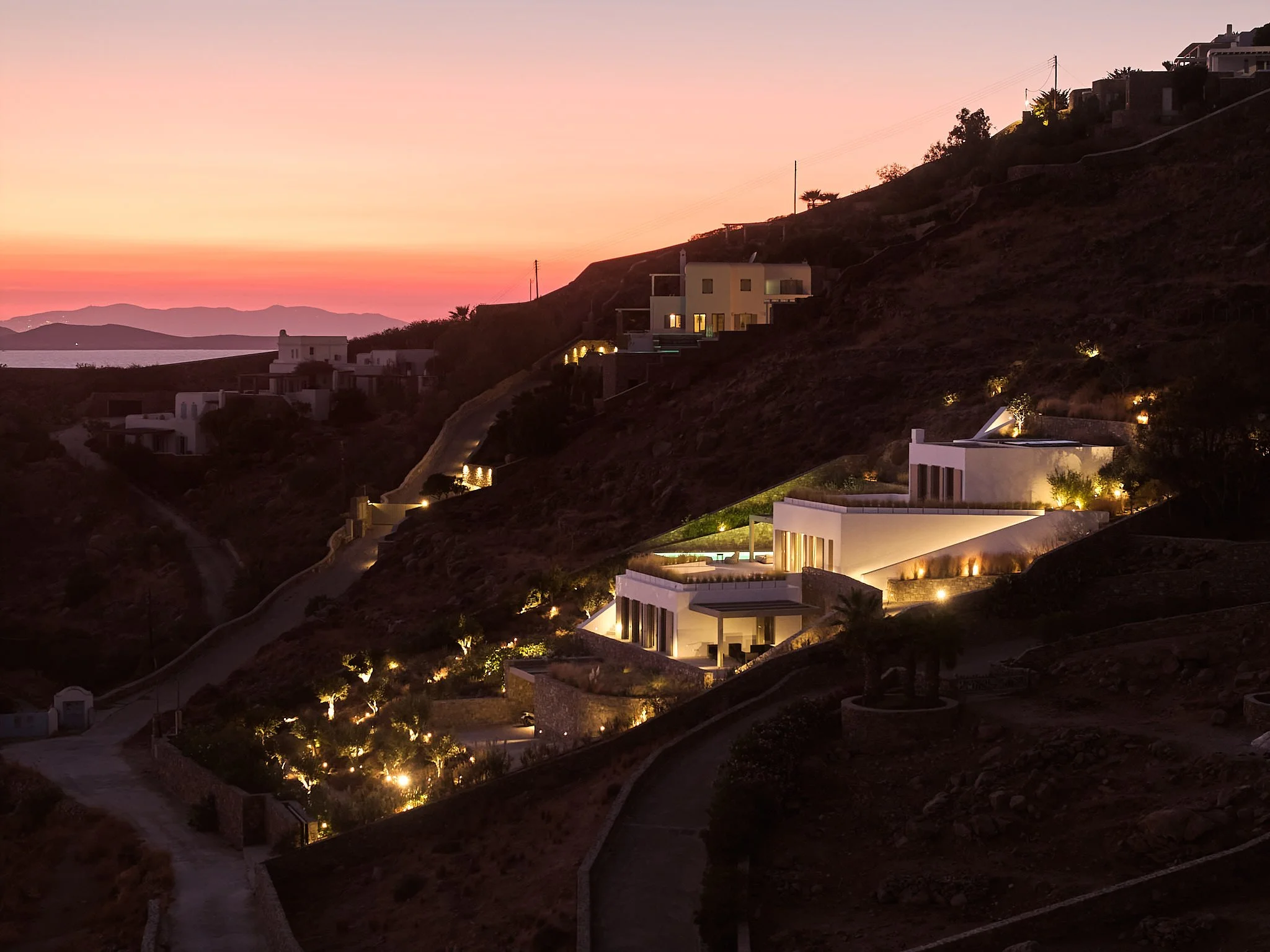 Modern white houses on a hillside at sunset with pink and purple sky, illuminated by outdoor lights.
