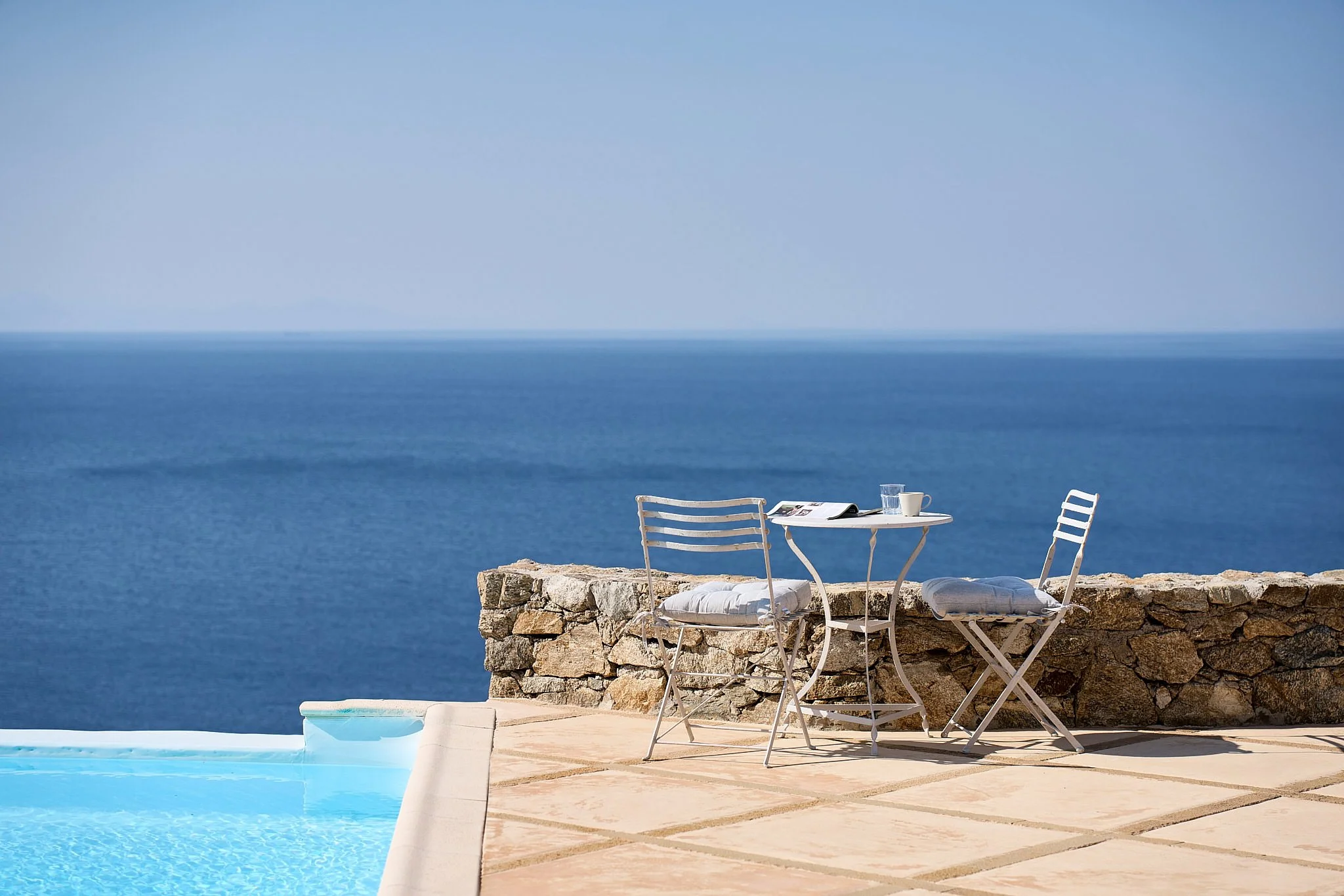 Poolside patio with white metal table and two chairs, overlooking blue ocean horizon, with a stone wall in the background.
