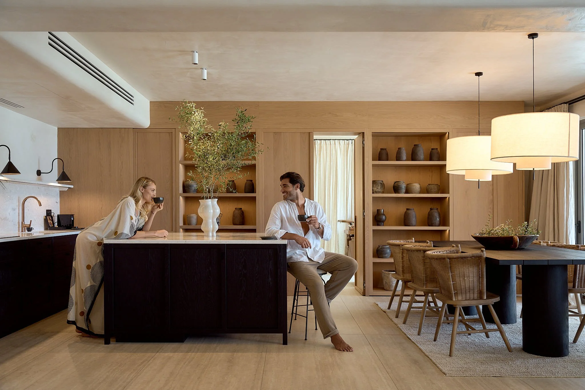 A man and woman in a modern kitchen having coffee and chatting, with a large island, wooden shelves with pottery, and warm lighting.