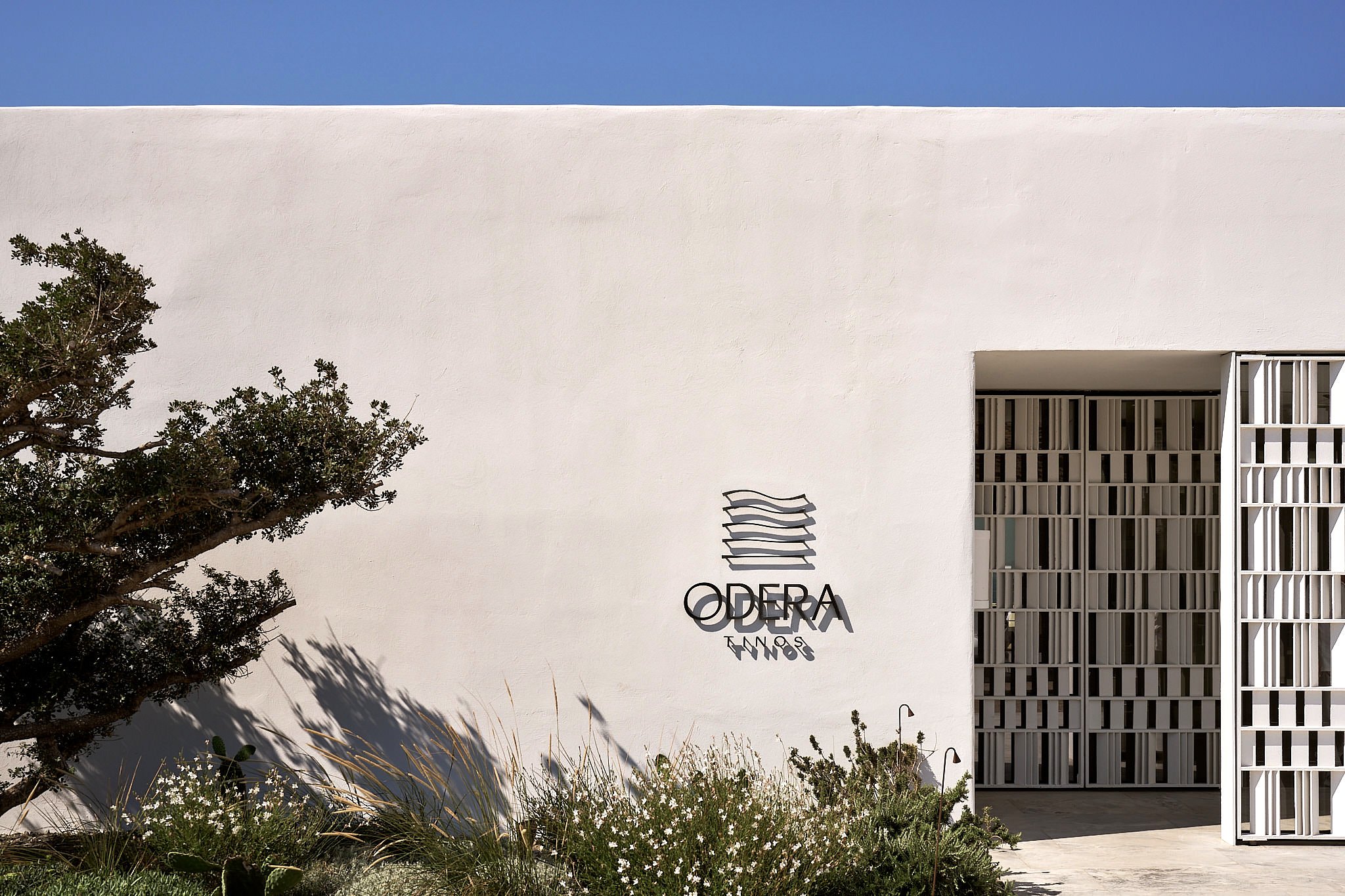 Modern white building with the sign 'Odera Tinos' and a stylized logo; a tree and plants in the foreground, blue sky overhead.