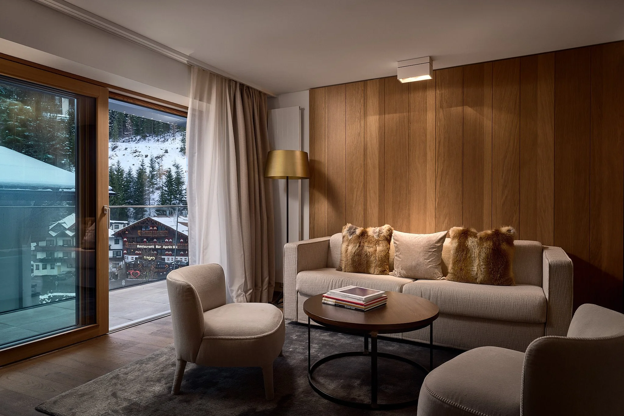 Interior of a modern living room with a beige sofa, furry throw pillows, a round wooden coffee table with magazines, two beige armchairs, a wooden accent wall, a floor lamp, and an open sliding glass door showing a snowy mountain landscape with build