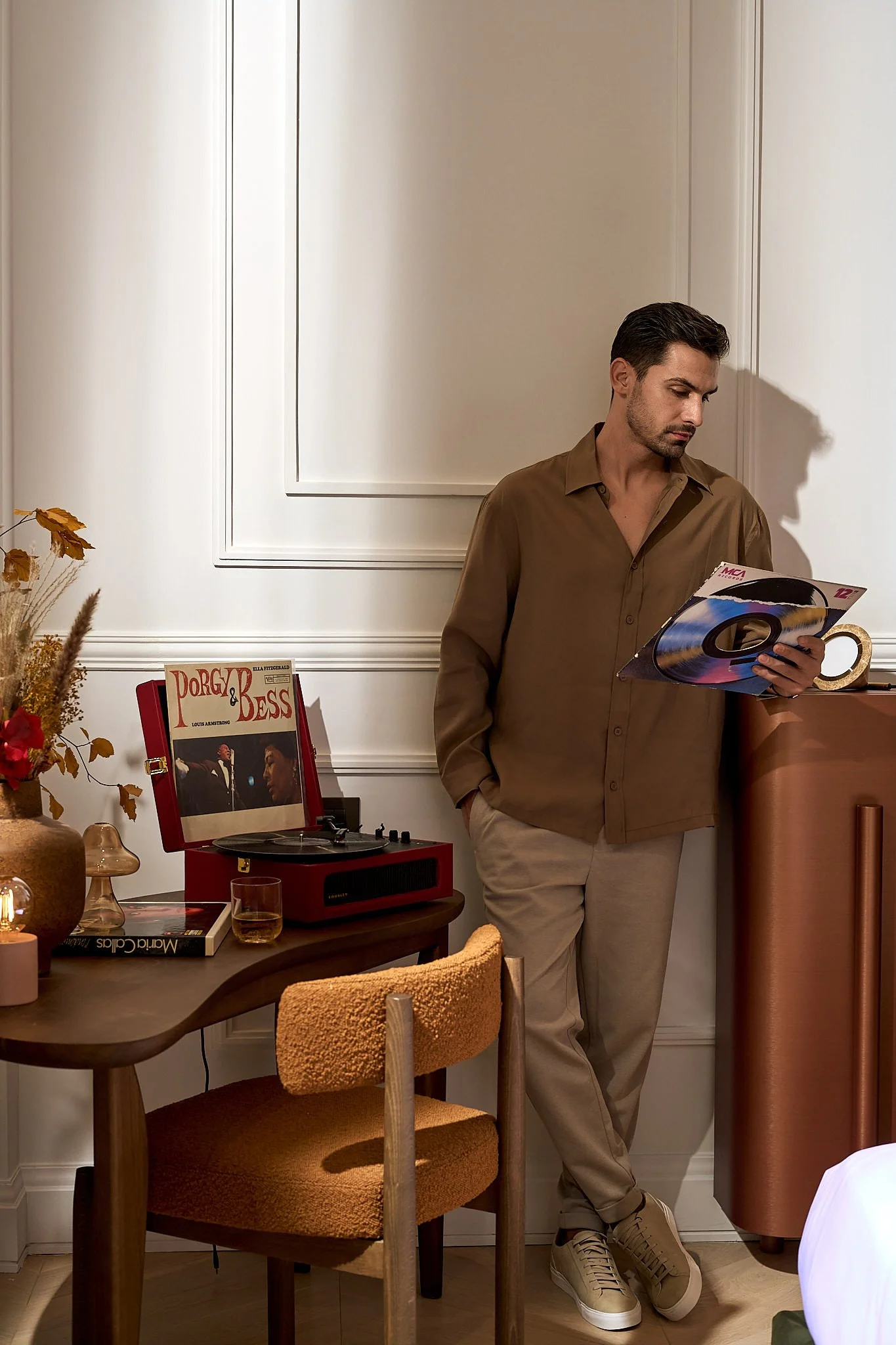 A man in a brown shirt and beige pants standing by a round wooden table, looking at a vinyl record in a sleeve. The table has a vintage record player, a glass of whiskey, and books. The wall behind has decorative molding, and there's a dresser with a
