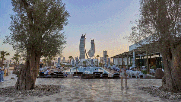 A waterfront area with boats docked and a city skyline featuring a distinctive twin-tower building in the background. People are walking and relaxing on the paved pathway flanked by large trees.