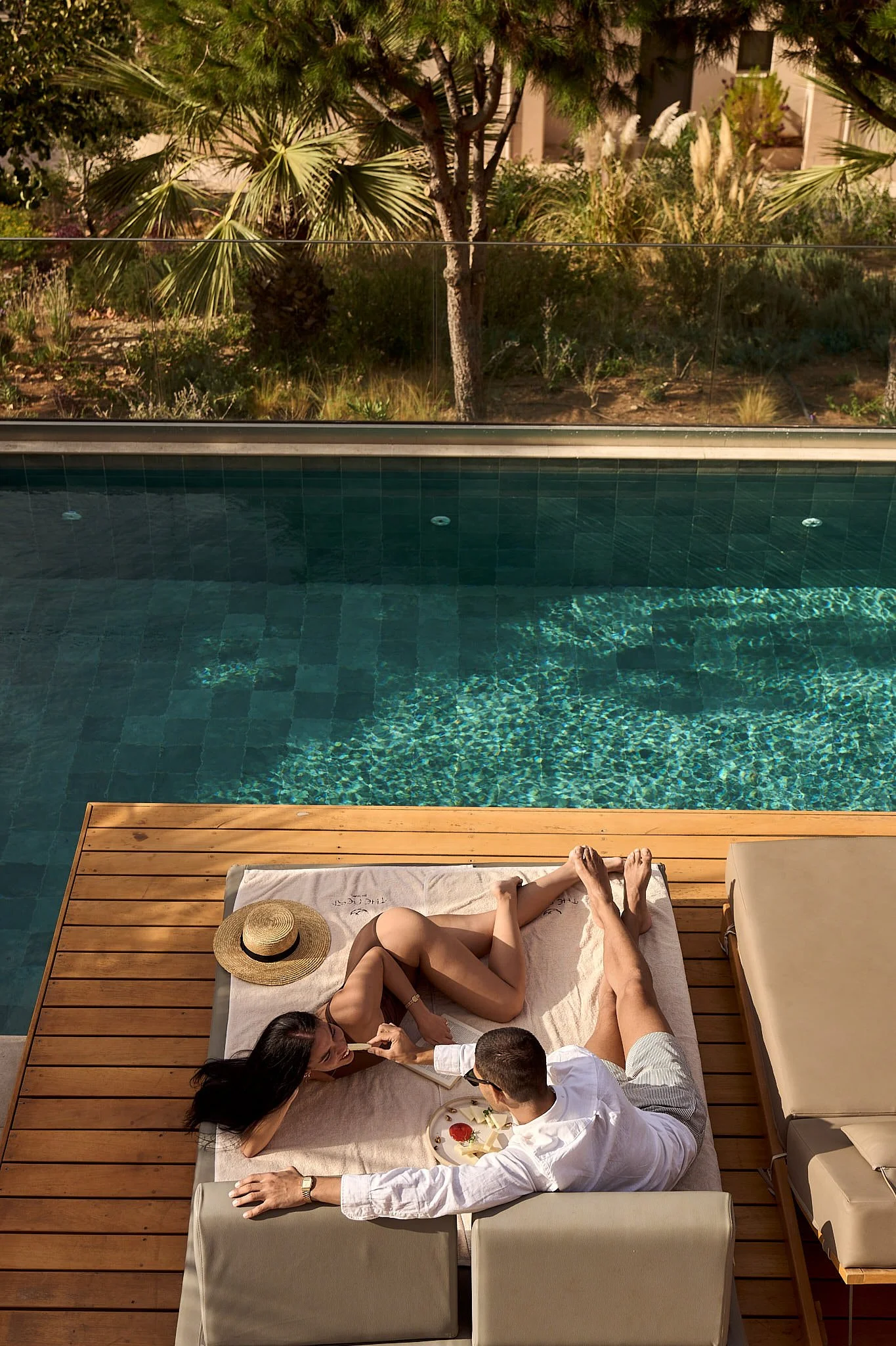 A couple relaxing on a towel-covered lounge chair by a swimming pool, with a straw hat beside them, having a snack.