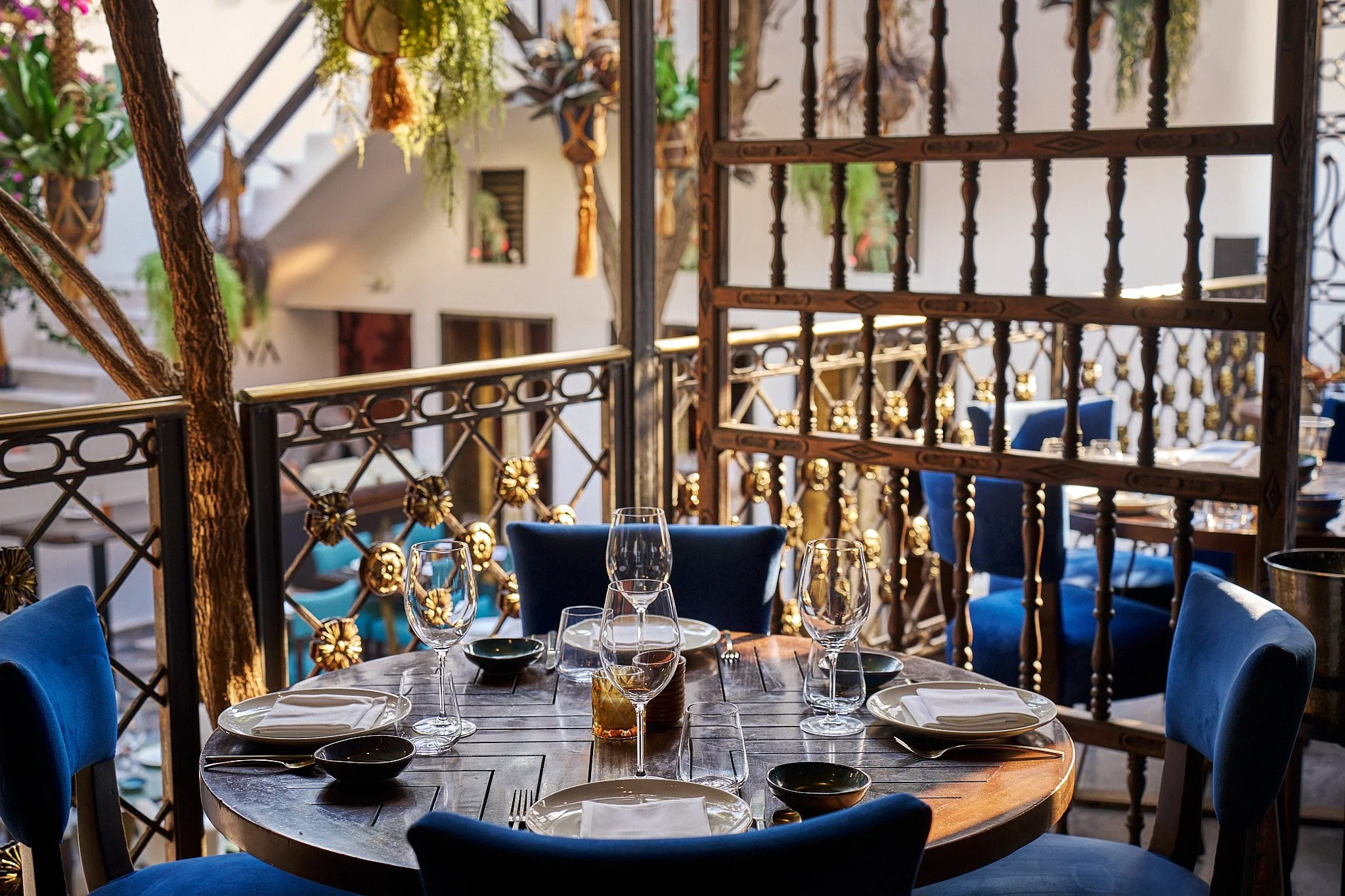 A round restaurant table set with glasses, bowls, and plates, surrounded by blue velvet chairs, on an outdoor balcony with decorative railings and hanging plants.