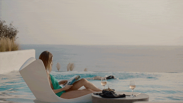 Woman relaxing by an infinity pool overlooking the ocean, reading a book with two glasses of wine nearby.