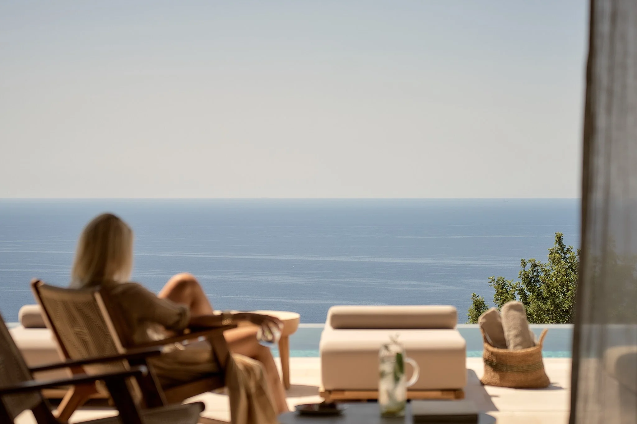 A woman relaxing on a lounge chair on a patio overlooking the ocean with clear blue sky, surrounded by outdoor furniture and greenery.