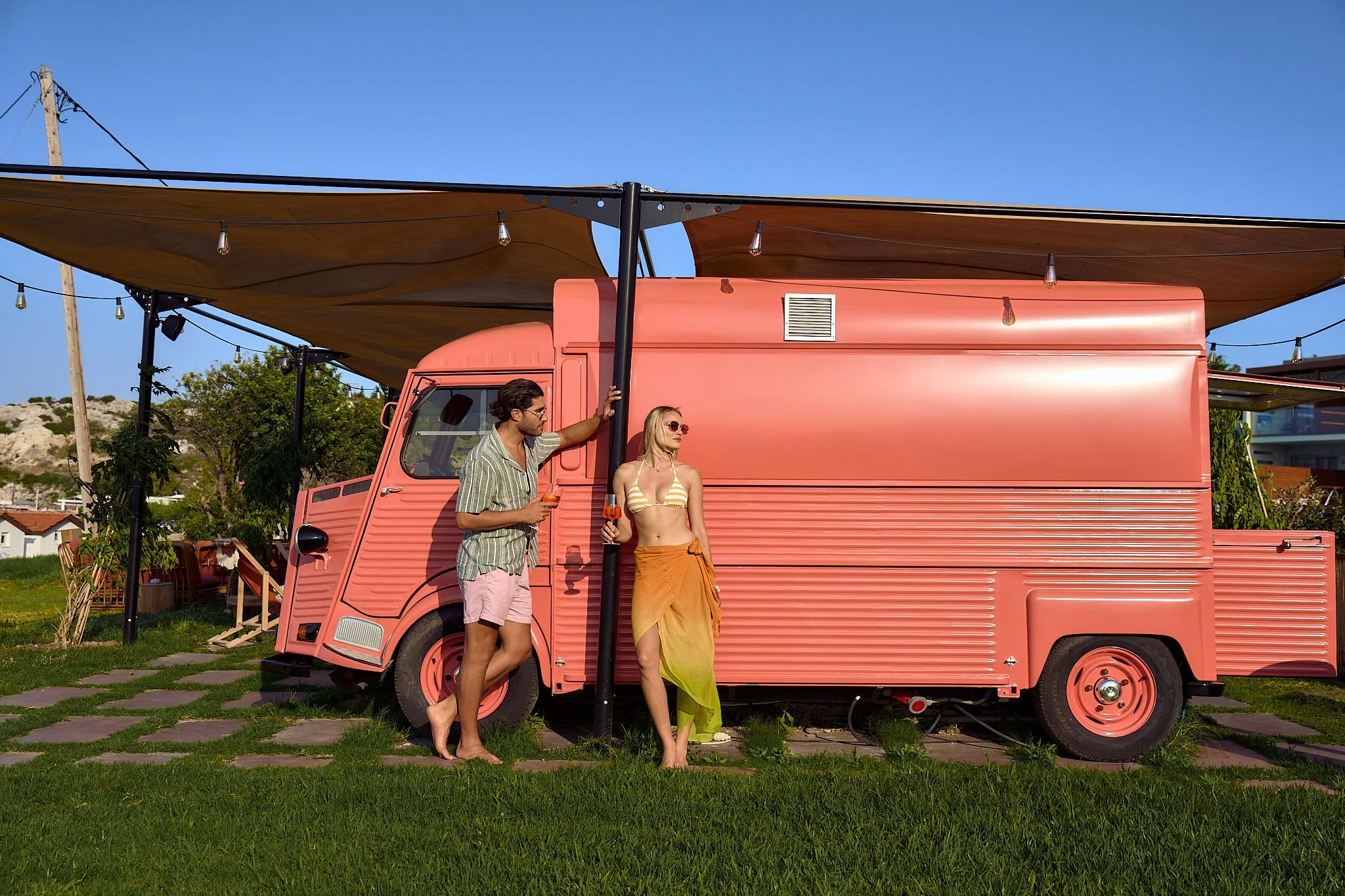 A pink vintage camper van with an awning, parked on grass with two people relaxing nearby under clear blue skies.