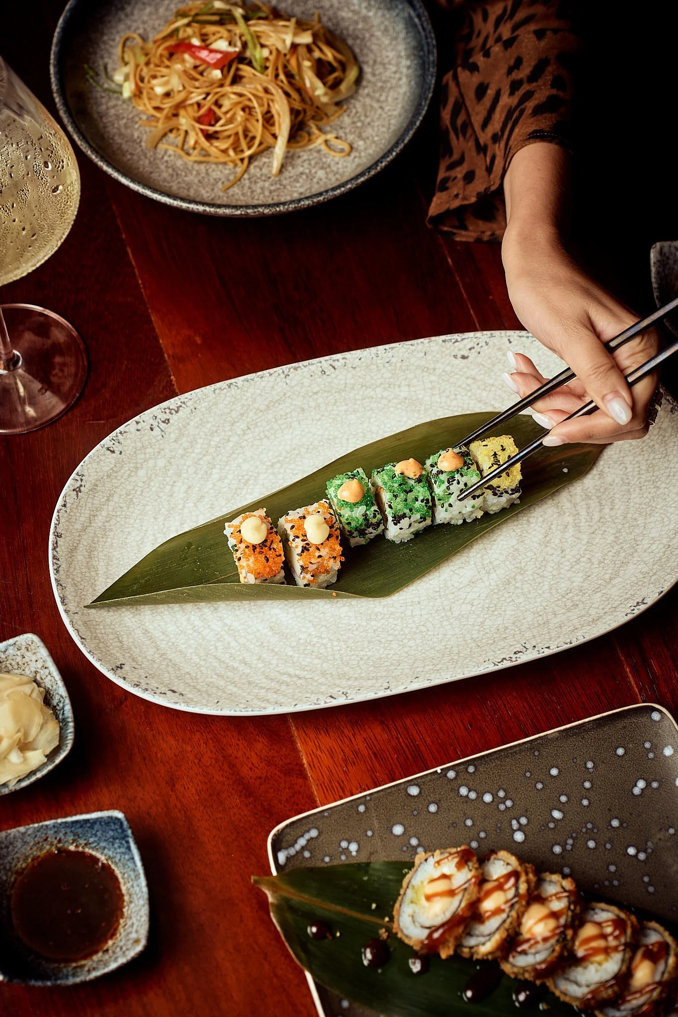 Assorted sushi rolls on a plate with a leaf, chopsticks, and side dishes, on a reddish-brown wooden table.