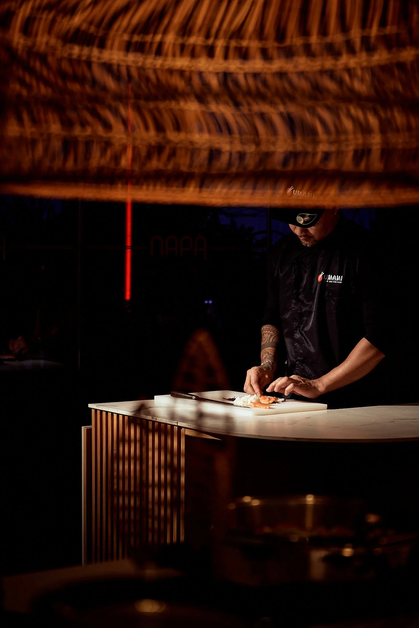 A man with tattoos and a black cap, wearing a black jacket with a logo, is preparing sushi at a sushi bar with a white countertop, in a dimly lit environment.