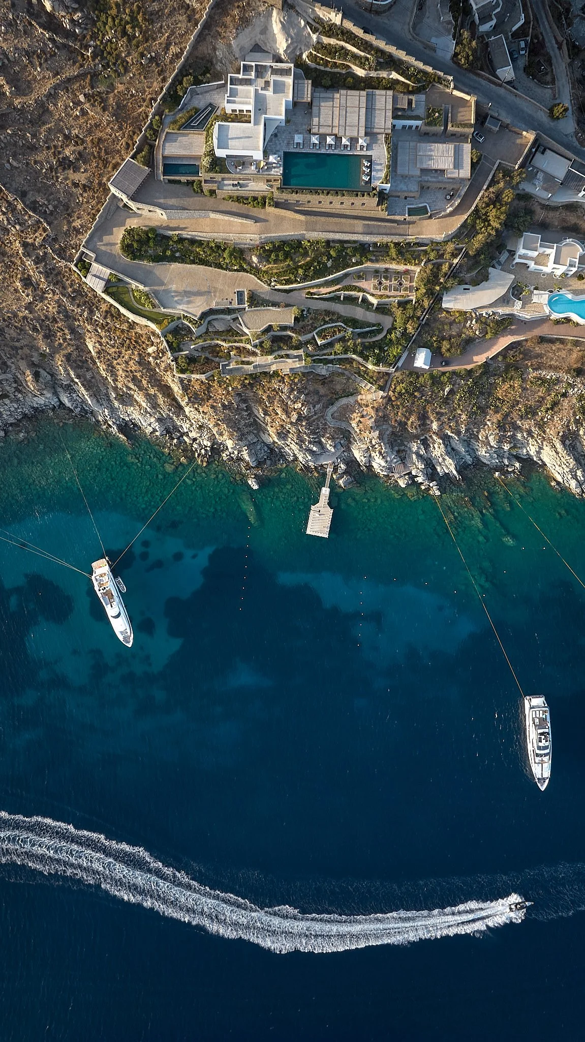 An aerial view of a luxury cliffside property with a large swimming pool, multiple buildings, landscaped gardens, and pathways, overlooking the ocean with boats docked nearby.