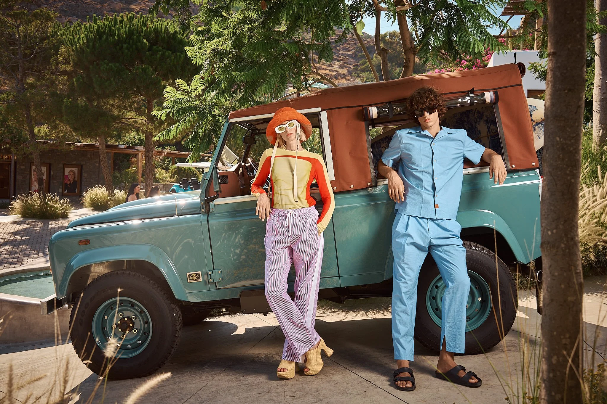 Two people leaning against a vintage teal off-road vehicle with a brown soft top, parked outdoors surrounded by trees with mountains in the background. The person on the left is a woman with blonde hair, wearing a wide-brimmed orange hat, white sungl