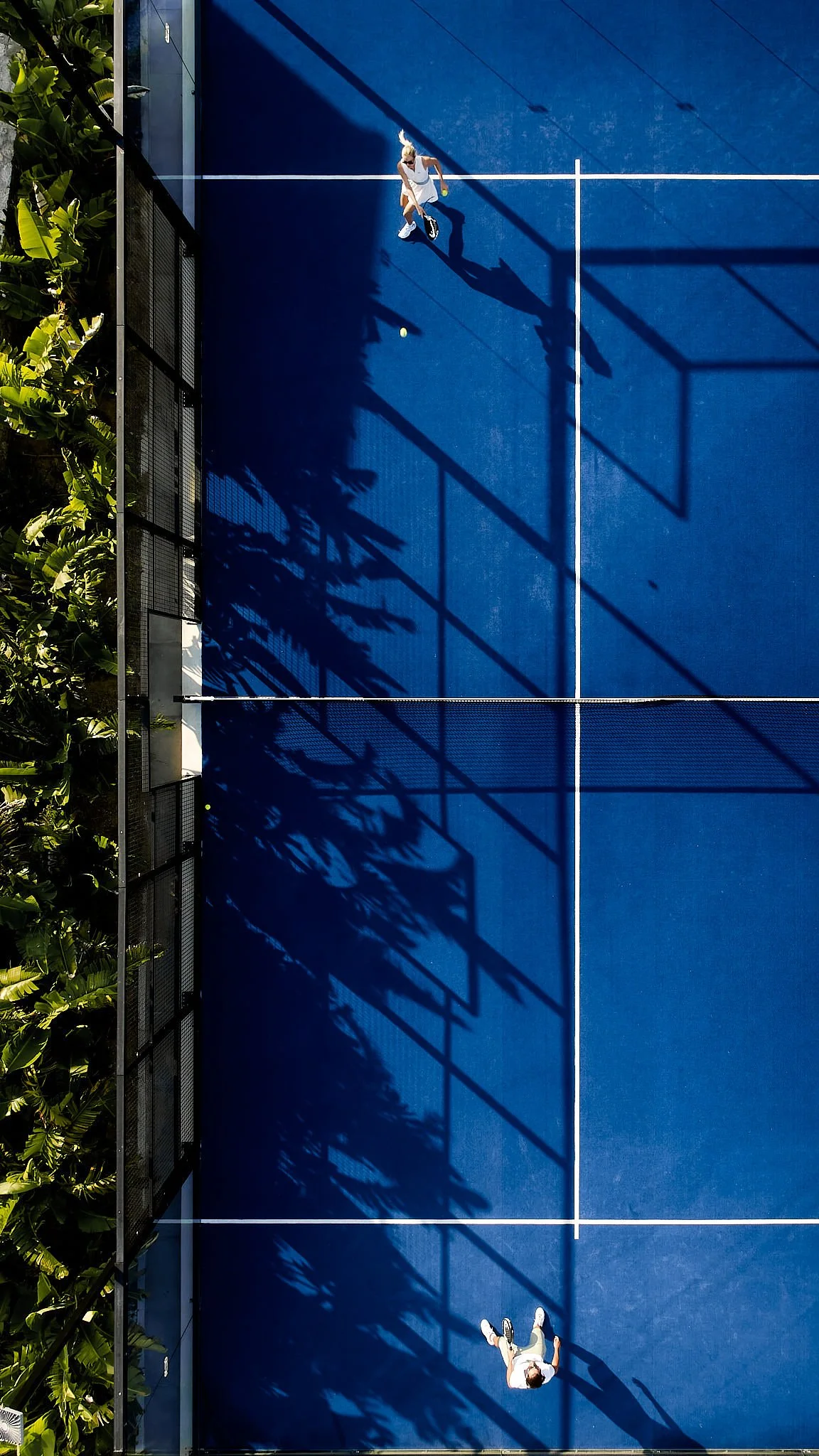 A bird's eye view of two people playing tennis on a blue court, one at the top serving and the other at the bottom preparing to return, with shadows cast by the court's fencing and surrounding trees.