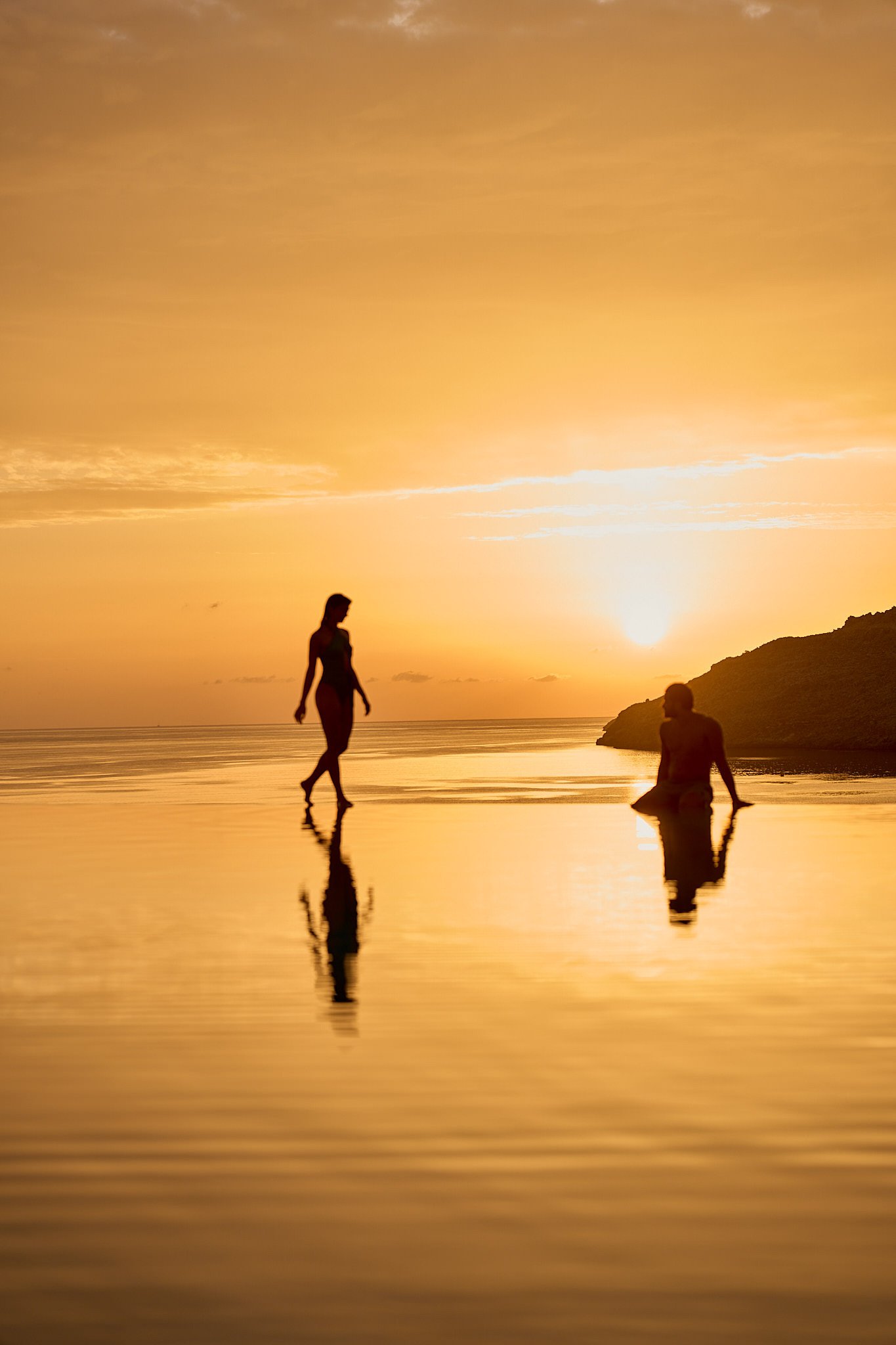 Silhouettes of two people on a beach at sunset, with reflections on water and a hill in the background.