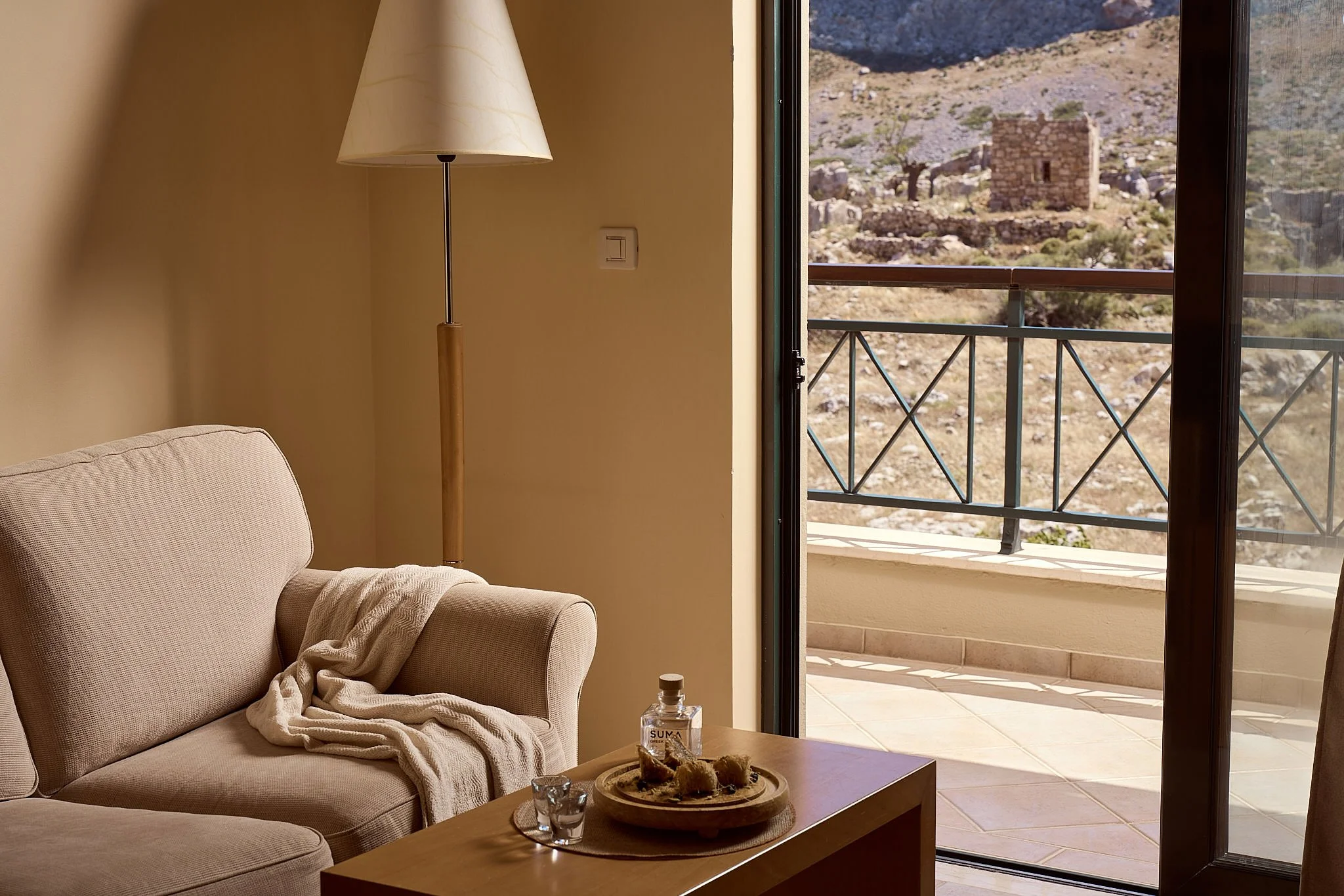 Living room with beige sofa and throw blanket, coffee table with snacks and a water bottle, open sliding glass door leading to a balcony with mountain view.