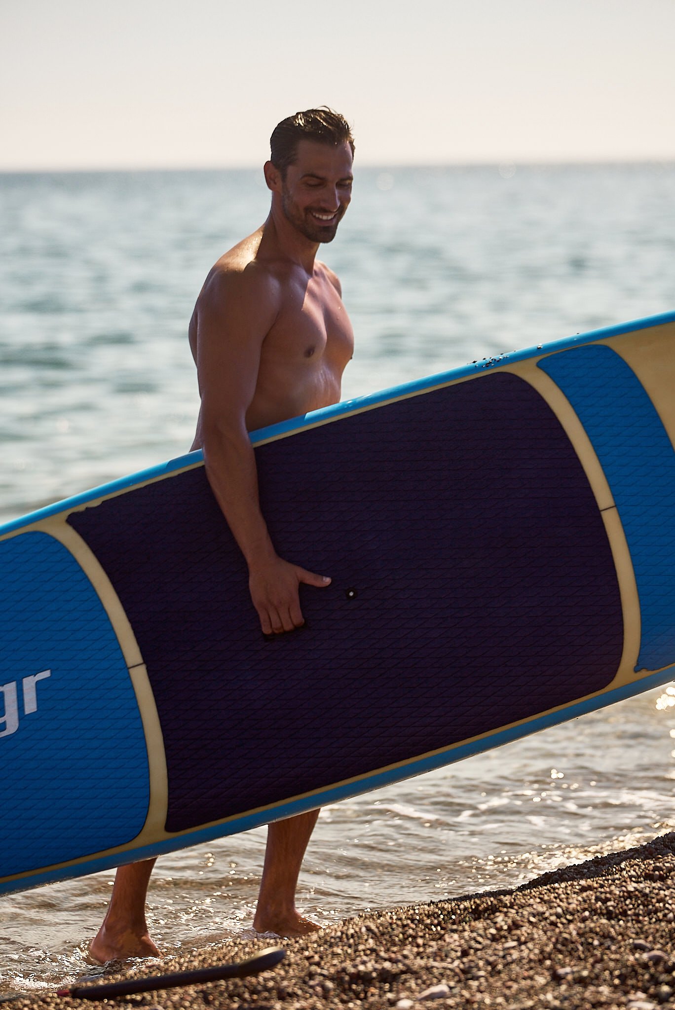 A man carrying a large blue and yellow stand-up paddleboard at the beach, smiling and looking down, with ocean water and a pebble shoreline in the background.