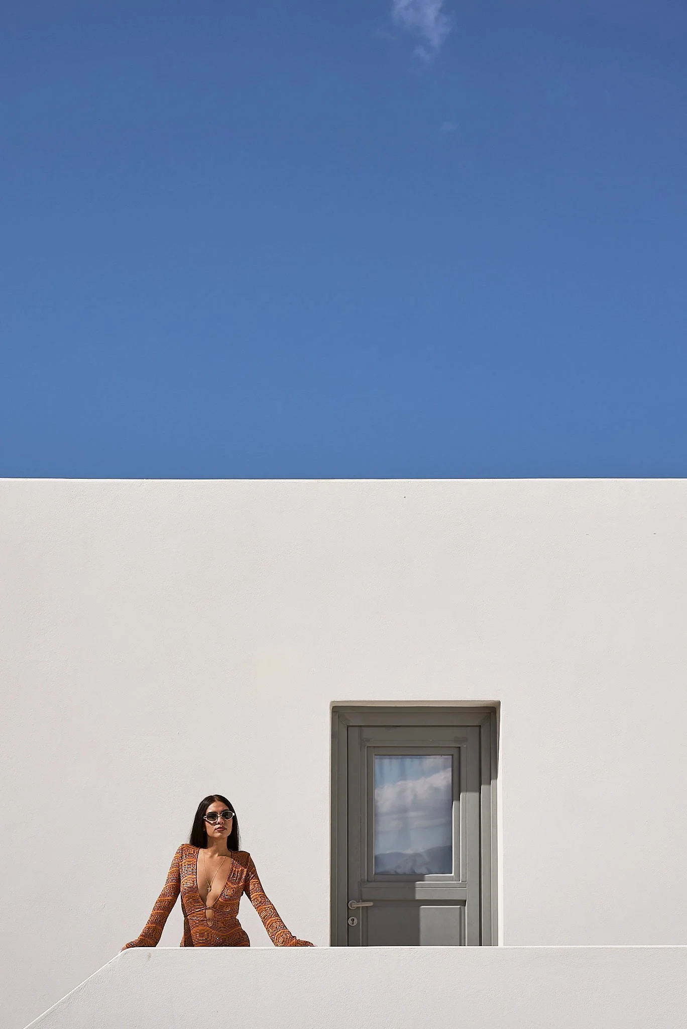 A woman wearing sunglasses and a patterned dress leaning on a white railing in front of a white wall with a gray door, under a blue sky.