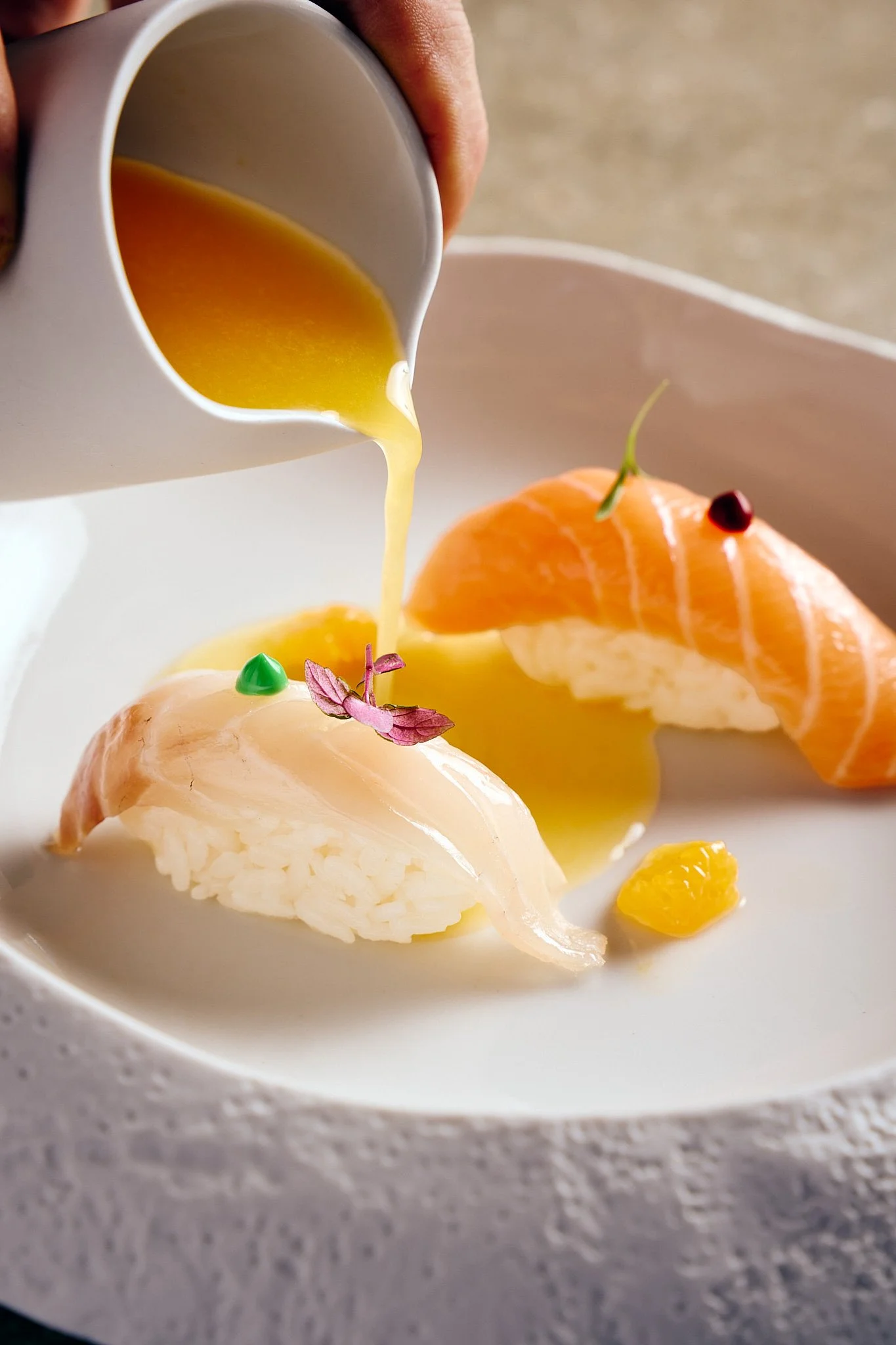 A plate with sushi, including slices of raw fish (salmon and white fish) on rice, garnished with tiny edible flowers and microgreens, with yellow sauce being poured over the sushi.