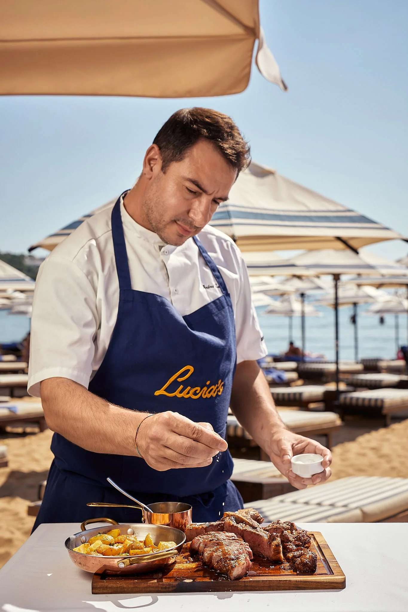 A chef is preparing grilled steak and roasted potatoes at a beachside restaurant under umbrellas.