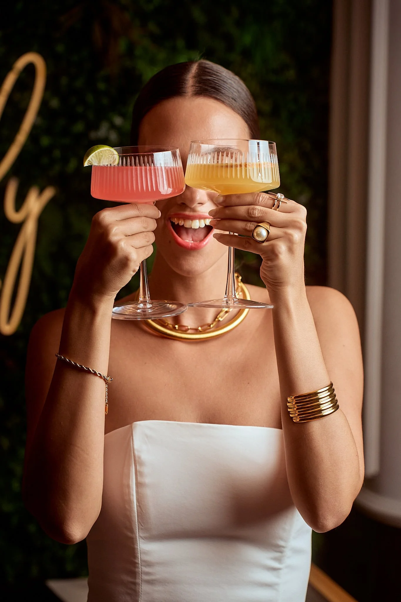 A woman with dark brown hair smiling with open mouth, holding two cocktail glasses in front of her face, one with a pink drink and lime wedge, the other with a yellow drink, in a celebratory setting with greenery background.