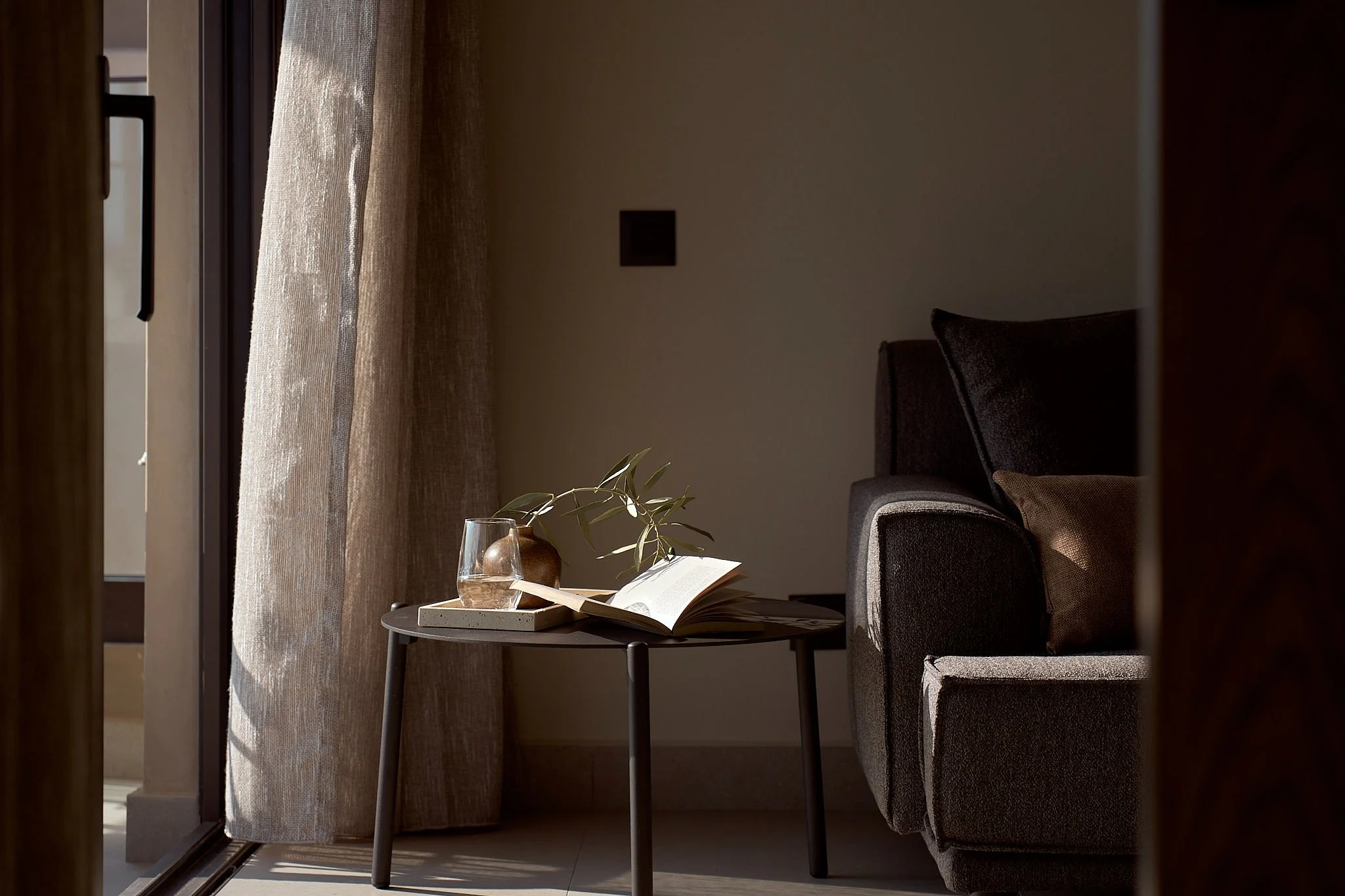 Living room corner with a small black table holding an open book, a glass vase with a branch, and a coaster. A gray armchair is partially visible on the right, next to a wall with a switch. Sunlight filters through a sheer curtain on the left.